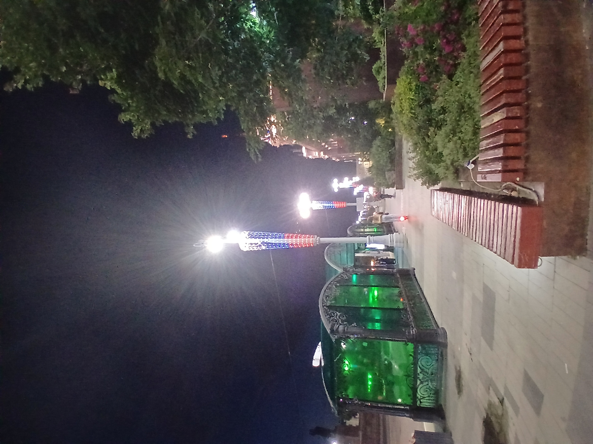 This image shows a night scene on a pedestrian street in Yekaterinburg, Russia. The foreground features two long, red-brown wooden benches with a darker base. To the left, a structure with green glass panels and ornate black metal railings is visible. Beyond this, a lamppost adorned with red, white, and blue lights stands prominently, emitting a bright glow. Further down the street, another similar lamppost is visible, along with people riding scooters. The background is dominated by the dark night sky and the silhouette of a building with illuminated windows. A large tree with dark green foliage is on the right side of the frame. The street appears to be paved with light-colored tiles.