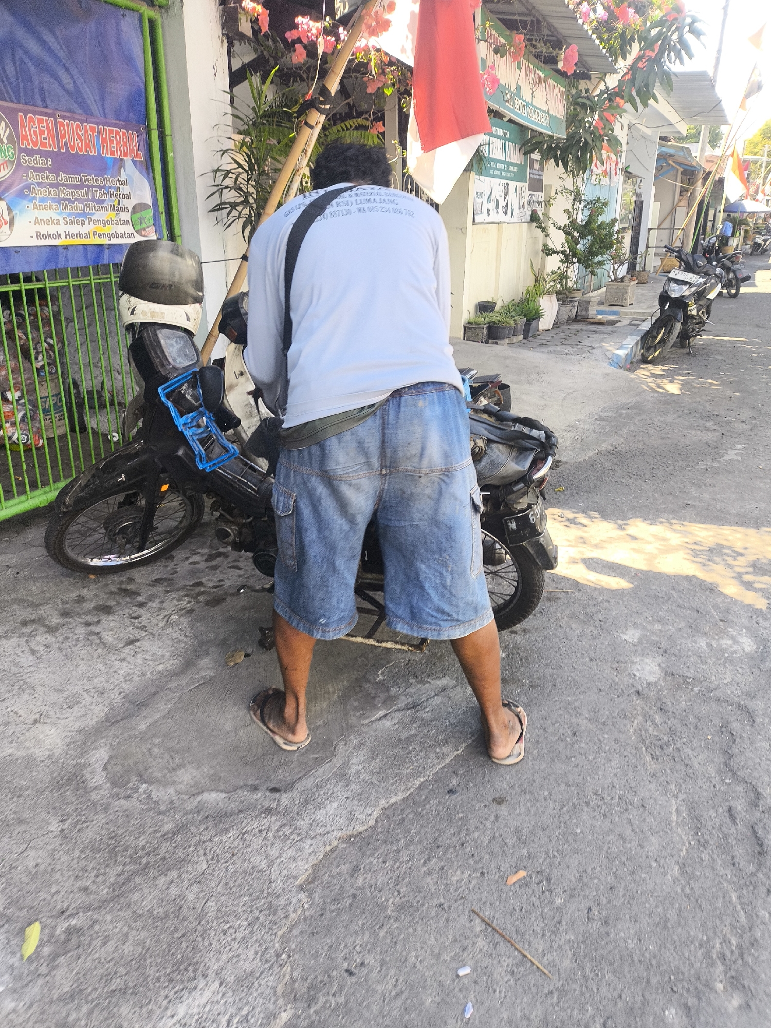 The image depicts a street scene in Lumajang, Indonesia. A person wearing a light-colored long-sleeved shirt and denim shorts stands with their back to the camera, facing a parked motorcycle. The motorcycle is dark-colored with a blue accessory attached to the handlebars. To the left of the motorcycle, a green metal fence encloses items that appear to be discarded plastic bottles and bags. Behind the fence, a blue banner with text in Indonesian is visible, including "AGEN PUSAT HERBAL" which translates to "Herbal Center Agent." Several potted plants are arranged along the wall to the right of the motorcycle. In the background, other motorcycles are parked, and a building with red and white flags is visible, suggesting a local business or event. The ground is paved with concrete, and sunlight casts shadows across the scene.