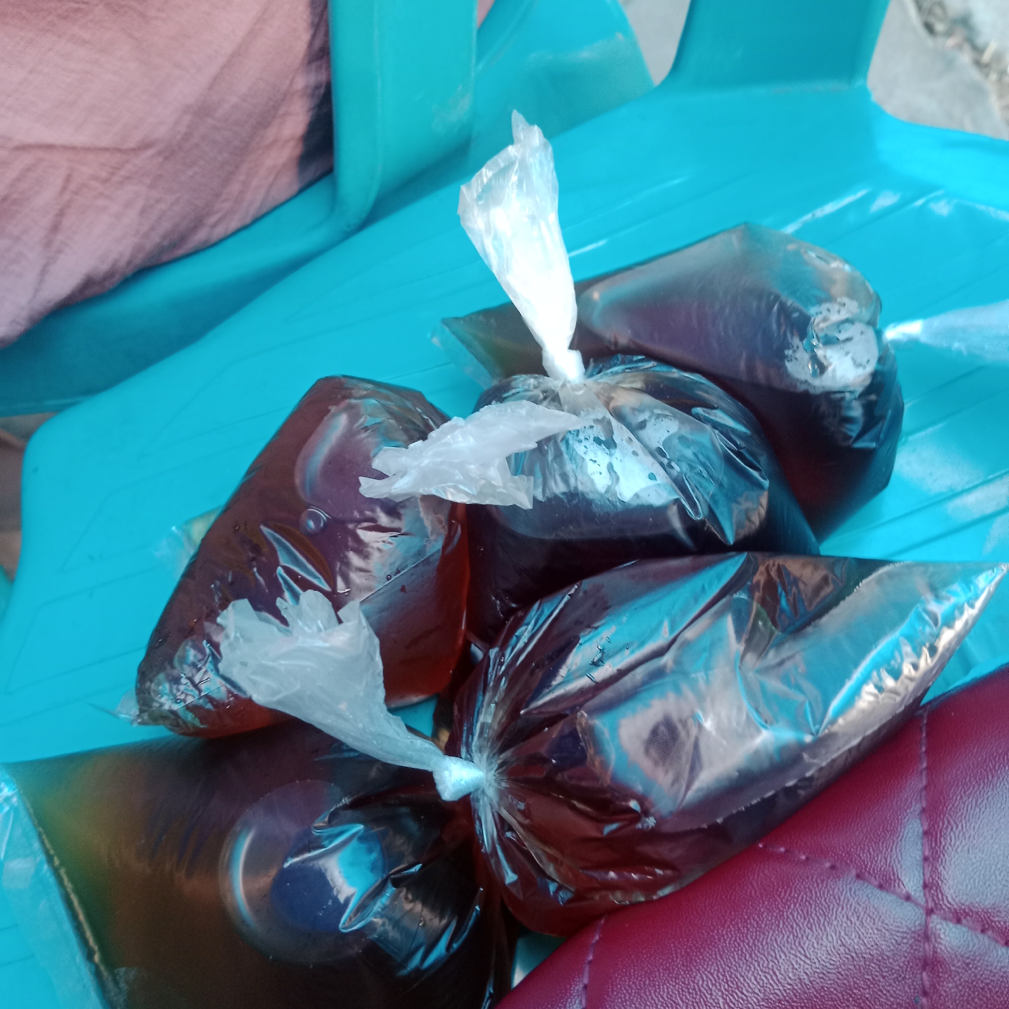 The image shows four plastic bags filled with a dark liquid, likely a beverage. The bags are tied with white plastic ties. They are placed on a bright turquoise surface, possibly a plastic chair or table. A portion of a person wearing a pink garment is visible in the upper left corner, and a section of a dark red quilted surface, possibly a seat cushion, is in the lower right corner. The scene is brightly lit, with reflections visible on the plastic bags. The overall context suggests an outdoor or informal setting. The image is taken from a slightly elevated angle, looking down at the bags. The image is consistent with a scene in Tegowanu Kulon, Indonesia, where such beverages sold in plastic bags are common.