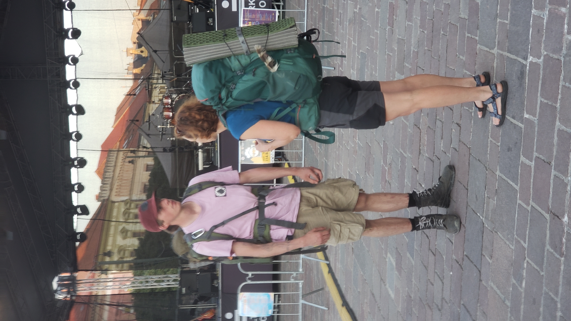 The image captures two individuals standing on a cobblestone plaza in front of an outdoor stage setup. A young man on the left, wearing a maroon baseball cap, a pink t-shirt, khaki cargo shorts, black socks, and dark hiking boots, is facing a woman on the right. The man has a large green backpack strapped to his shoulders. The woman, with short, curly reddish-brown hair, is wearing a blue t-shirt, dark athletic shorts, and dark sandals. She has a large green backpack with a rolled-up sleeping pad attached to the back, also on her shoulders. The background shows scaffolding, stage lighting, and what appears to be a large screen or backdrop with an image of buildings. The plaza is partially visible, with a yellow line marking a boundary on the left side of the image. This scene is consistent with an outdoor event or festival taking place in the city of Prešov, Slovakia, likely in its historic center given the architectural style of the buildings in the backdrop.