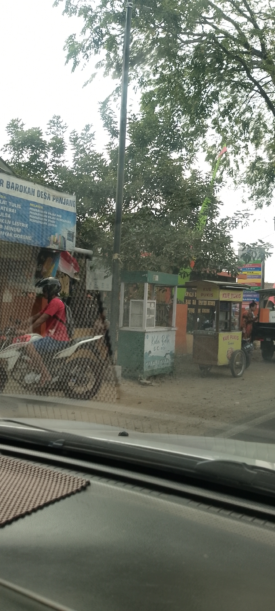 The image depicts a street scene in Panjang, Indonesia, with a focus on a roadside stall and passing traffic. In the foreground, a dark dashboard of a vehicle is visible, suggesting the photo was taken from inside a car. A person on a motorcycle, wearing a red shirt and a helmet, is moving past the scene from left to right. The motorcycle has a white and black body. Behind the motorcycle, a blue banner is partially visible with text in Indonesian, including "BAROKAH DESA PANJANG," indicating the location. The banner also lists services like "MELAYANI PEMBAYARAN PAJAK PBB," "PEMBAYARAN PAJAK KENDARAAN," and "PEMBAYARAN LISTRIK," suggesting it's a service point.
Adjacent to the banner, there are several small roadside stalls. One stall has a green base and a white counter with the text "KEDAI KELA," and "SCANTI." Another stall, slightly to the right, is yellow and advertises "KUE PUKIS." A third stall, to the right of the yellow one, also advertises "KUE PUKIS." A woman in an orange and brown patterned dress is standing at the rear of a dark truck behind the stalls. The environment appears to be dusty, with dirt and some debris on the ground. Trees with dense green foliage are visible in the background, and a utility pole with a wire runs vertically through the scene. The sky is overcast and bright. The image captures a typical Indonesian street scene with local businesses and daily life.