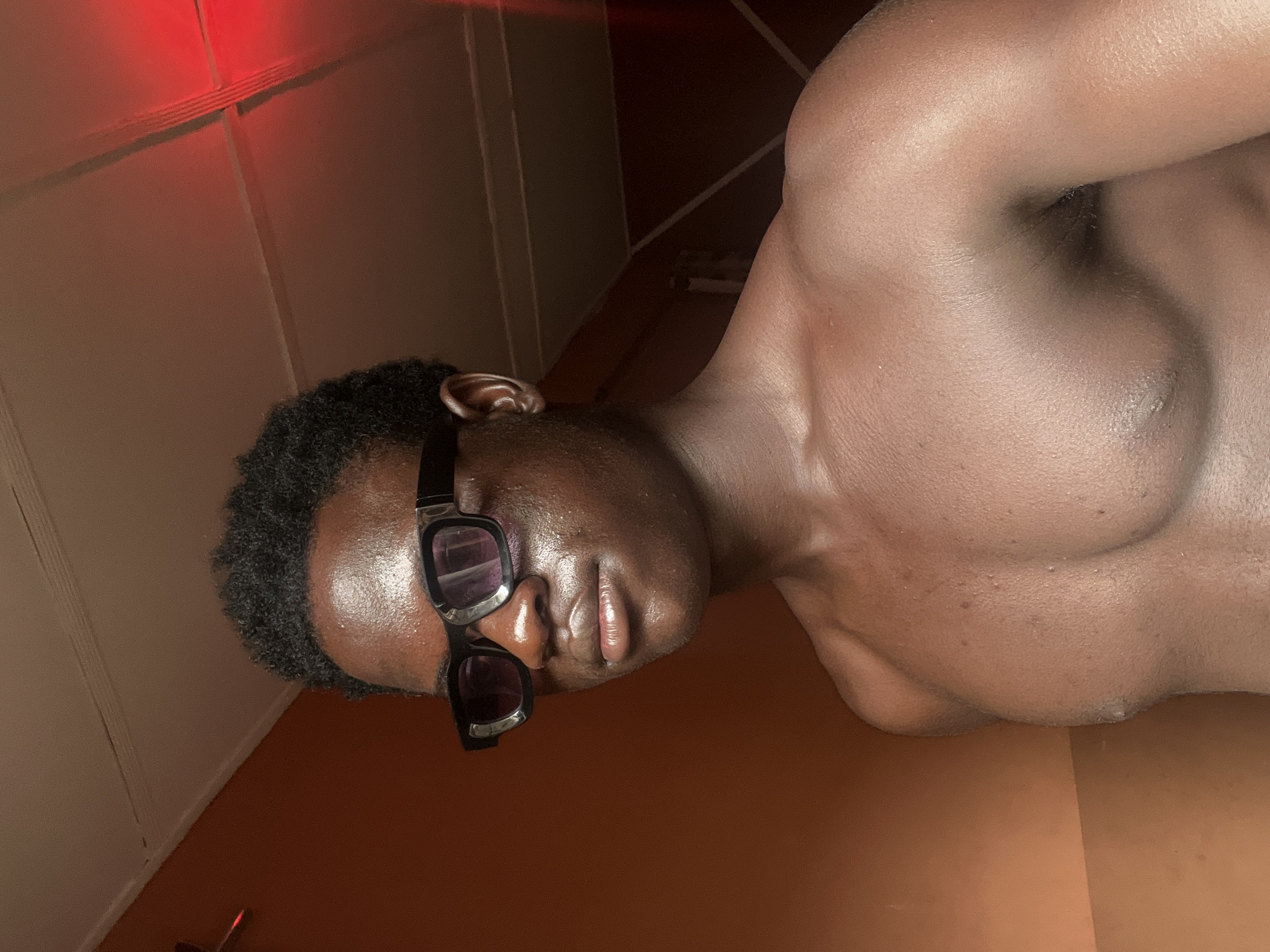 A tanned, muscular man with sunglasses reclines. He has short, dark hair and the sunglasses are black with dark lenses. He appears oiled or sweaty. The background consists of a wood-paneled wall with a red light source at the top left and a slanted ceiling. The floor is brown. There is no specific evidence to indicate the location is Mosafejaw, Nigeria.