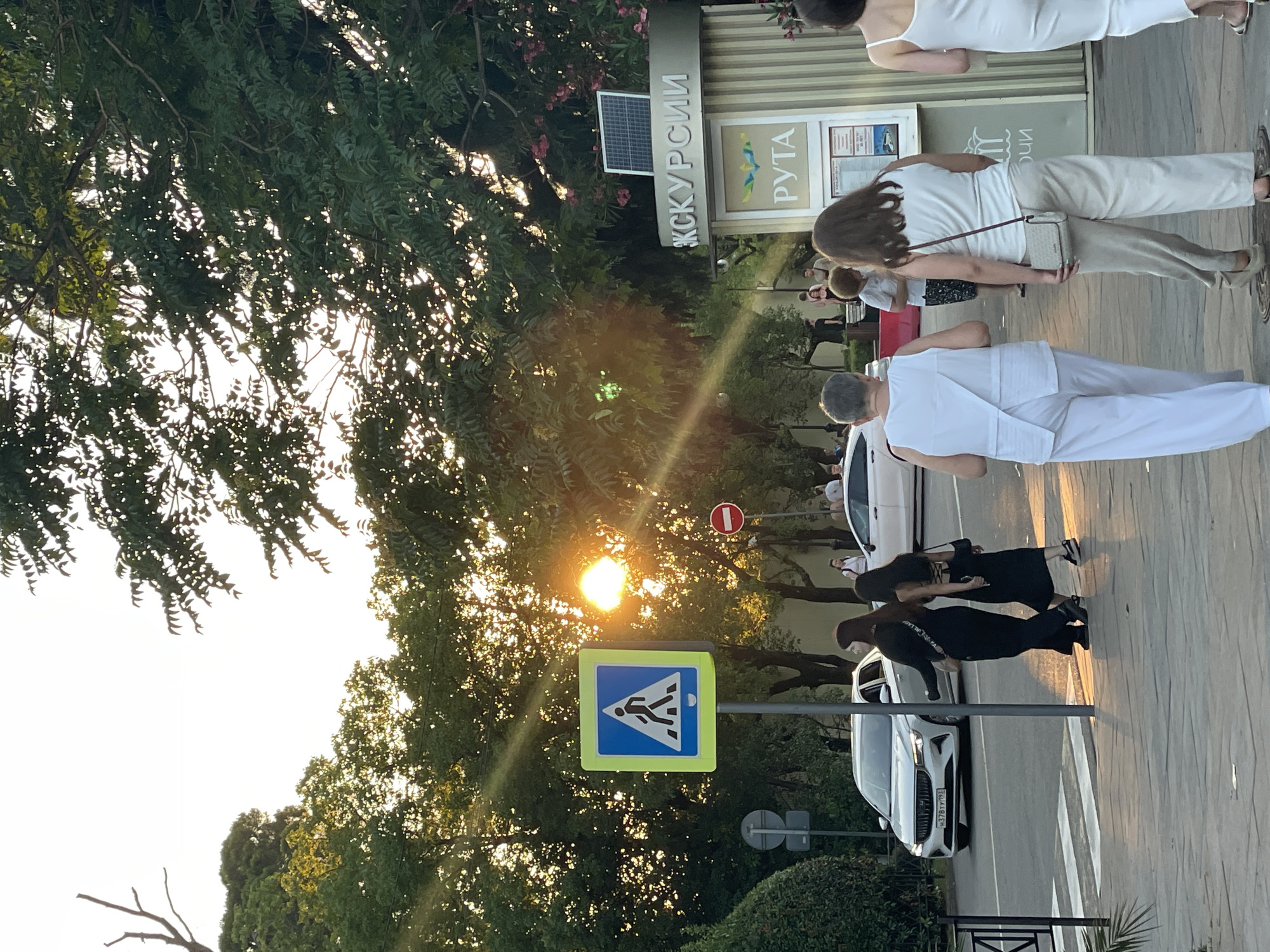 The image captures a street scene in Sochi, Russia, during the golden hour, with the sun low in the sky and casting long shadows. In the foreground, a pedestrian crossing is marked with white stripes on the asphalt. A prominent blue and yellow pedestrian crossing sign is visible on the left.
Several people are walking on the sidewalk. Two women in black dresses are walking away from the camera, with one carrying a light-colored crossbody bag. Further ahead, three people in white clothing are walking towards the camera: an older woman with short gray hair, a woman with dark brown hair wearing a white tank top and light beige pants, and a woman in a white sleeveless dress.
On the right side of the image, there is a kiosk with signage that includes Cyrillic text and the word "РУТА" (RUTA). Above this, another sign reads "ЭКСКУРСИИ" (Excursions). A small solar panel is mounted on top of the kiosk.
Cars are visible on the street, including a white sedan with a license plate that appears to read "H 378TY 777 RUS". A round red and white "no entry" sign is on a pole in the background. Lush green trees and some flowering bushes frame the scene, with the sunlight filtering through the leaves. The overall impression is of a vibrant public space bathed in the warm light of sunset.