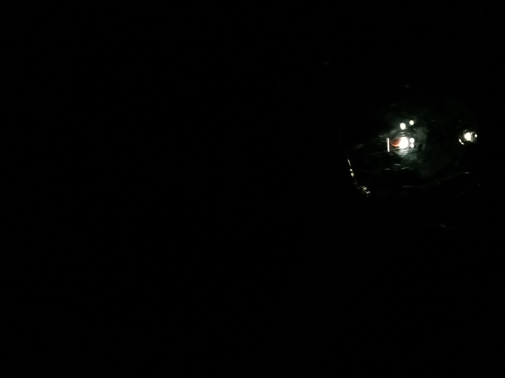 The image depicts a predominantly dark, almost entirely black scene, indicating nighttime or an extremely dim environment. In the upper right quadrant, various artificial light sources are visible as reflections on a dark, glossy, and somewhat textured or undulating surface.
The most prominent reflection is a reddish-orange rectangular light, positioned centrally within the illuminated area. Surrounding it are several smaller, circular or blob-like white lights, some appearing brighter and more defined than others. These lights are diffused and slightly blurred, suggesting either distance, motion, or reflections on an uneven surface. The reflective surface itself appears wet or highly polished, causing the light sources to stretch and distort slightly.
No people or distinct objects other than the reflections are visible. The lack of ambient light points strongly to it being night. The presence of clear reflections implies a wet environment, possibly a street after rain or a body of water. Given the location context of Lagos, Nigeria, this could easily represent reflections of city lights on a rain-slicked road or a puddle during the night. There are no discernible emotions or visible text in the image.