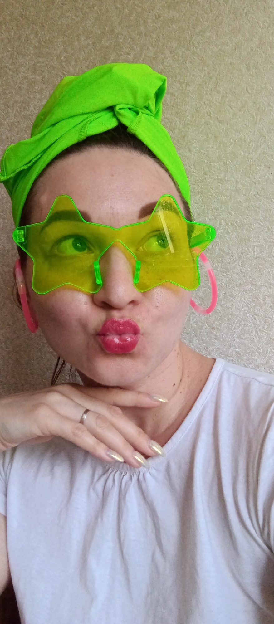 A woman with fair skin is posing for a selfie indoors. She is wearing a green headband, star-shaped sunglasses, and pink hoop earrings. Her lips are pursed as if she is blowing a kiss, and she is resting her chin on her right hand, which has long, pale yellow fingernails. She is wearing a white t-shirt. A silver ring is visible on her finger. The background is a neutral, textured wall. The city and location are unknown.