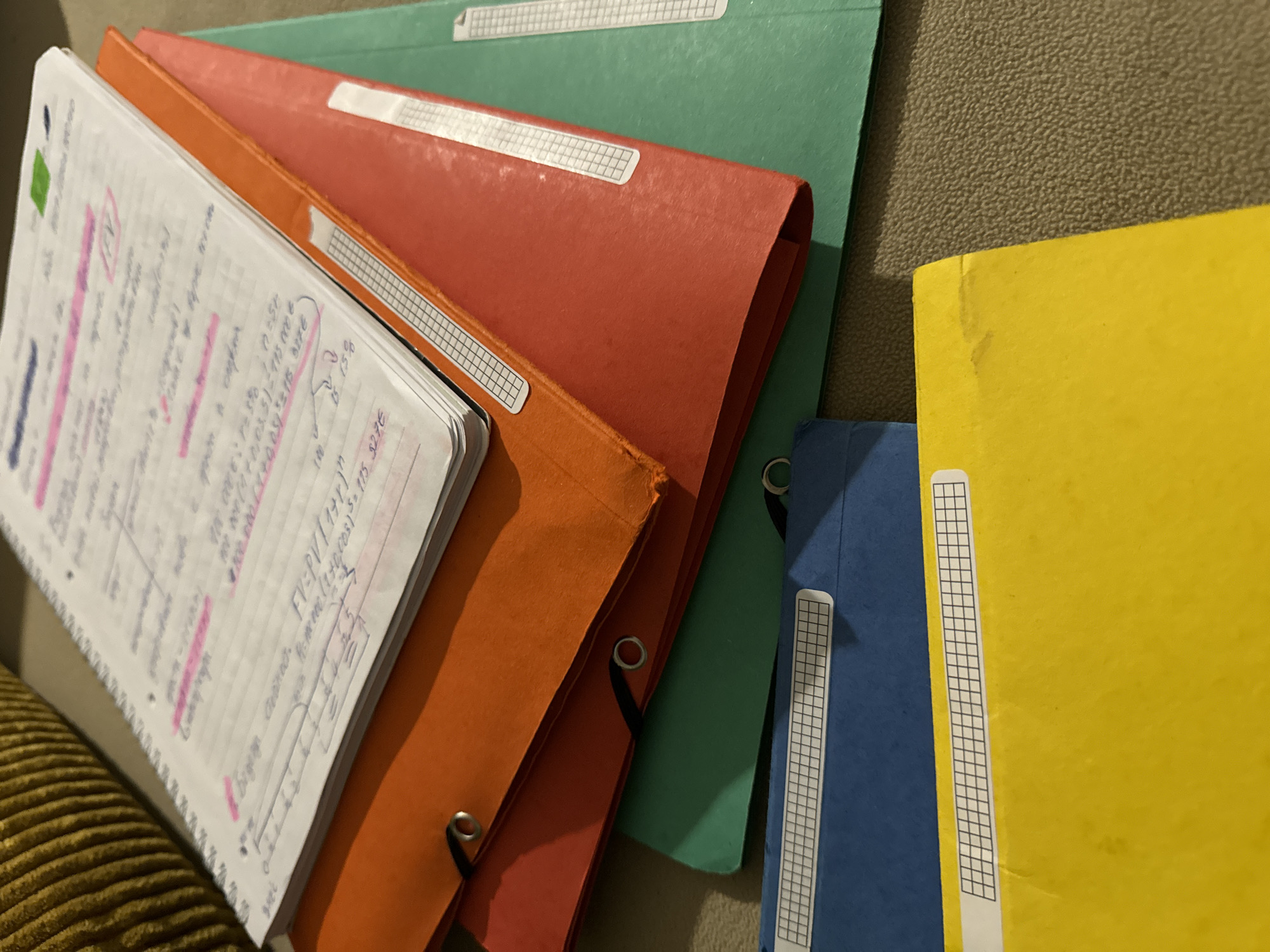 The image shows a close-up of several colorful ring binders stacked on top of each other. The binders are orange, red, green, blue, and yellow. A notebook with handwritten notes and markings is on top of the orange binder. The notebook has a white grid paper and is marked with pink highlighter. The text visible on the notebook contains mathematical equations and financial terms.