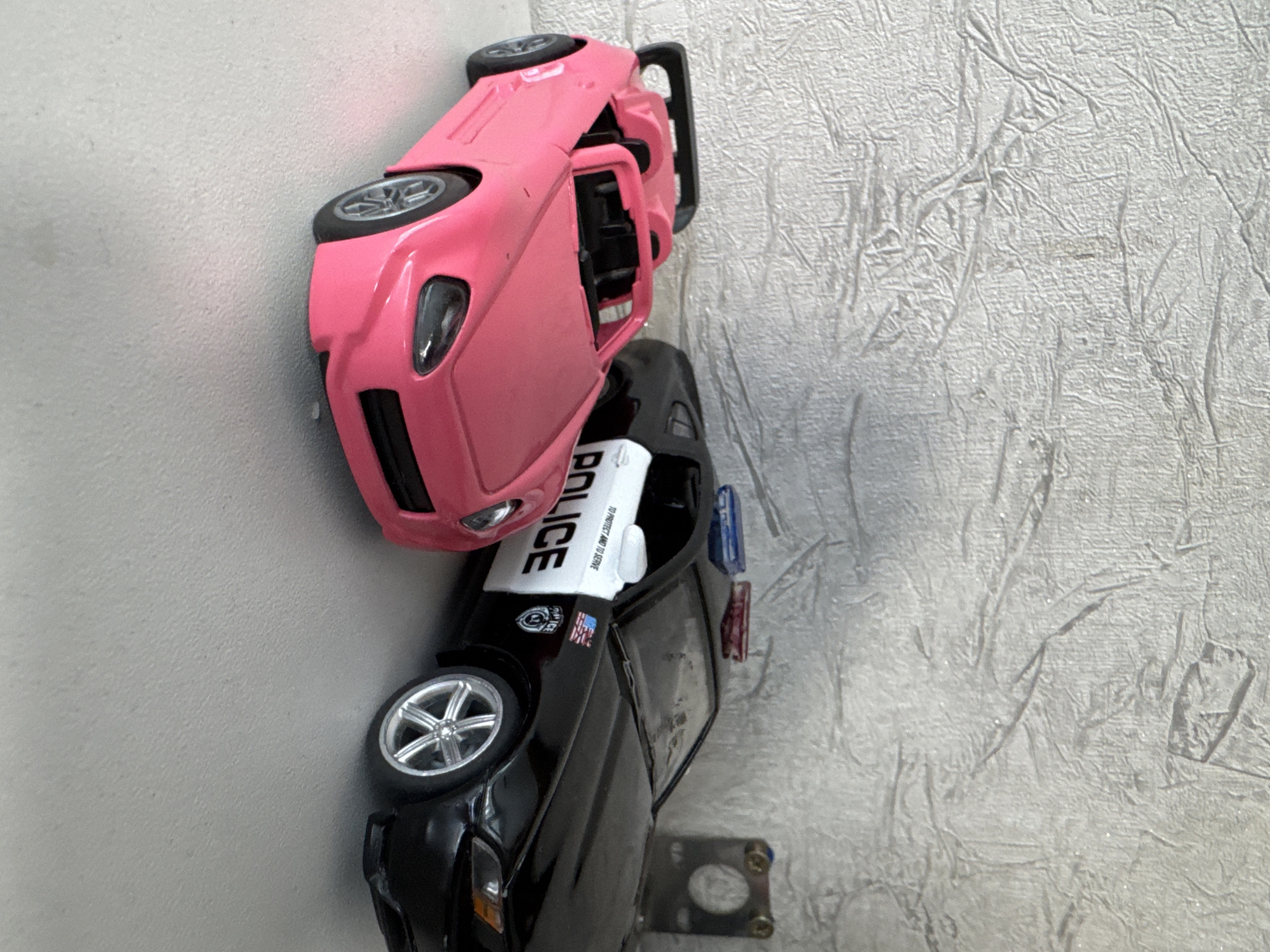 The image shows two toy cars placed on a white surface against a textured light-colored background. The car on the left is a pink convertible sports car. The car on the right is a black police car with "POLICE" written in white on its side, along with an American flag emblem and a shield. It also has blue and red lights on its roof. The overall scene appears to be a still life arrangement of toys. There is no indication of the location being in the city of Яромінскі сельскі Савет, Belarus.