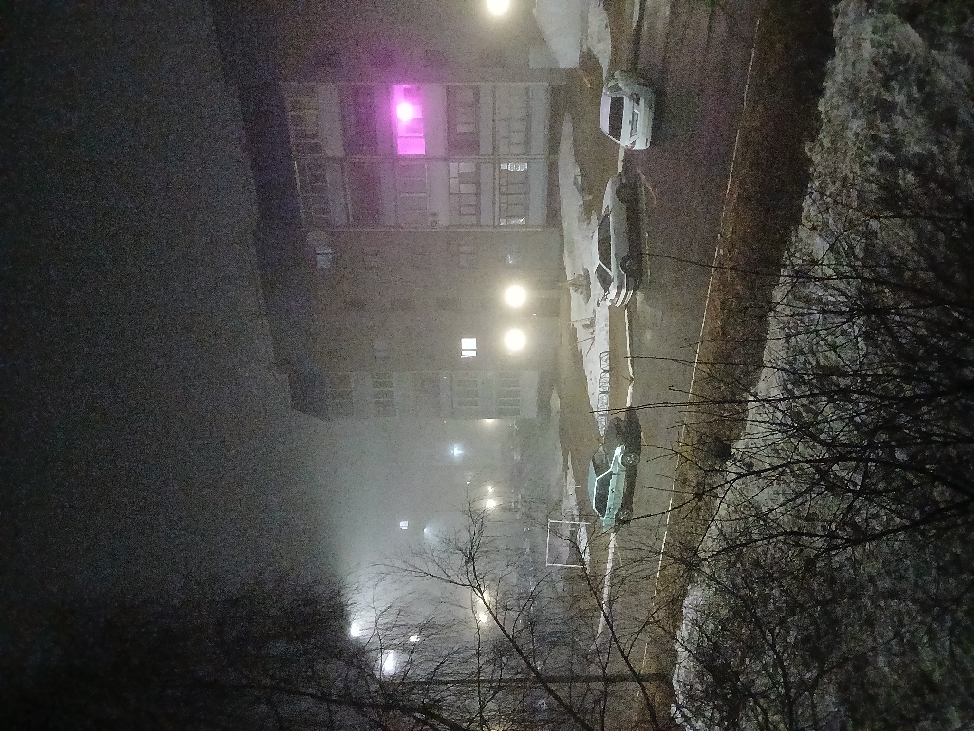 Nighttime view from an elevated position in Марковский, Russia, overlooking a street with parked cars, a building with a lit pink window, bare trees, and street lights. The atmosphere is foggy or misty.