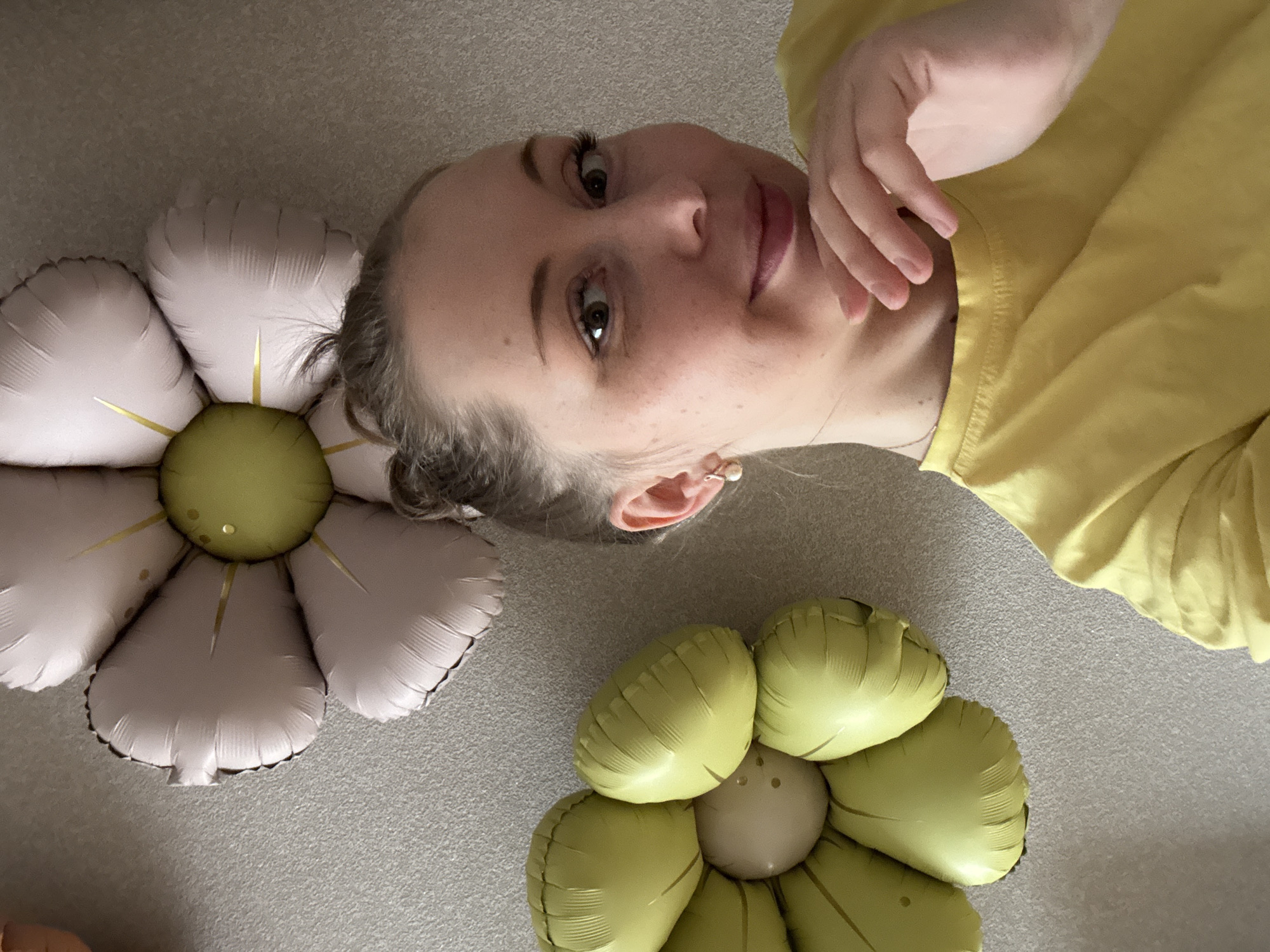 A fair-skinned woman with brown eyes and light brown hair pulled back in a bun is lying on a carpeted floor between two flower-shaped balloons. She is wearing a yellow shirt and has her right hand near her chin. The flower balloons are pink and green, and lime green and beige, respectively. The background is a light gray carpet.