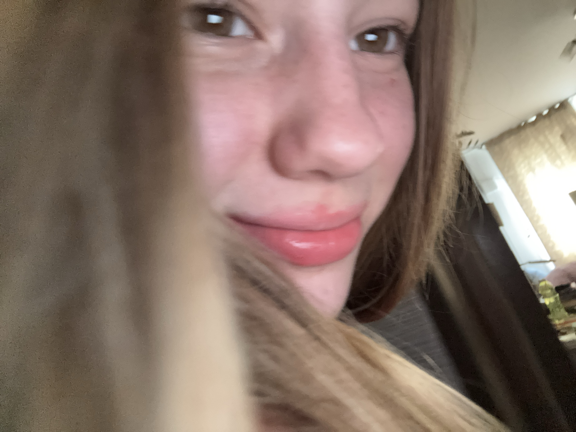The image is a close-up, slightly tilted selfie of a young person's face. The person has fair skin with noticeable pink undertones on their cheeks and nose, suggesting a flush or blush. Their eyes are brown with prominent eyelashes, and their eyebrows are light brown and well-defined. The nose is small and slightly upturned. The lips are full and appear to be coated with a glossy pink lip product. Strands of light brown hair are visible, obscuring parts of the face. The background is out of focus, but hints of what appears to be an indoor setting with wooden furniture and possibly a window with curtains can be discerned. There is no visible indication of the location within the city of Vsevolozhsk, Russia.