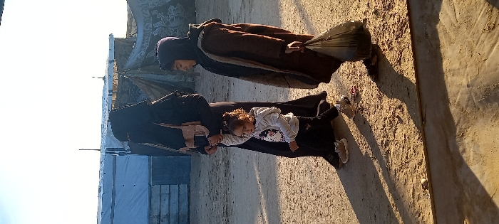 Three individuals walk across unpaved, sandy ground in Khan Yunis, Palestinian Territory, under a clear sky, with makeshift shelters constructed from tarps and other materials visible in the background. A woman on the left, dressed in a black abaya and niqab, carries a light brown shoulder bag and holds the raised right hand of a young child. This child, a girl with curly brown hair, wears a light grey long-sleeved top with a dark graphic, dark trousers, and light-colored sneakers, looking towards the left. To their right, an older child, dressed in a dark hooded garment and a long brown coat, carries a black plastic bag in their left hand, looking slightly downwards and to the right. All three individuals are moving from left to right across the frame, casting long shadows on the ground extending towards the left.