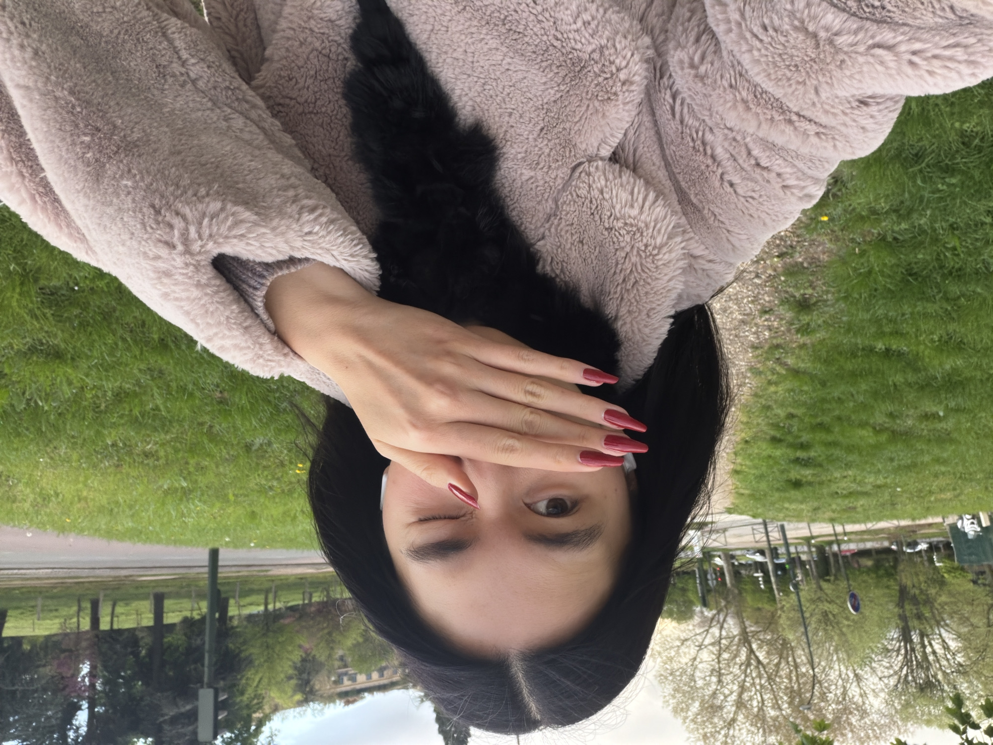 A young woman with long dark hair is winking at the camera while holding her hand over one eye. She has dark red fingernails and is wearing a plush, light-colored jacket with a black fur-like collar or detail. She appears to be outside in a park-like setting in Paris, France. In the background, there is grass, trees, and buildings that are recognizable as typical Parisian architecture. A pathway or road is visible, along with streetlights and other park-related elements. The image is rotated, as it should be oriented counterclockwise 90 degrees.
