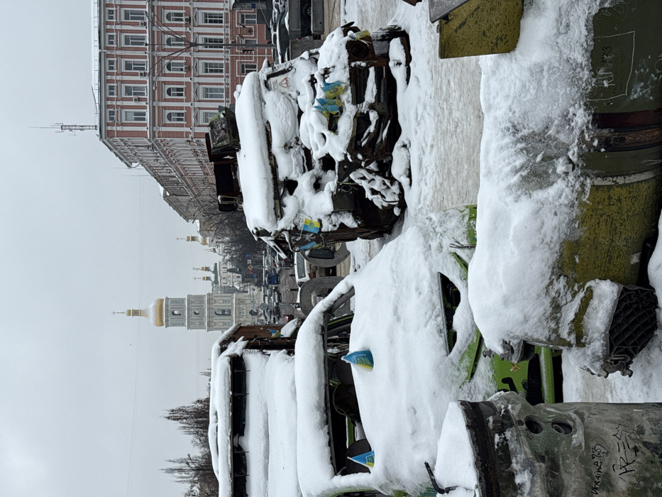 The image depicts an outdoor scene in Kyiv, Ukraine, during winter, with ground and objects covered in a layer of snow under an overcast sky. In the foreground and midground, three severely damaged, snow-covered objects are visible, consistent with war wreckage. To the left, a large, cylindrical, metallic object, possibly a missile or rocket casing, rests on its side; it bears faint graffiti including "Ukraine." Centrally, a heavily damaged green civilian car, likely a hatchback, is visible with its windshield missing; two small blue and yellow Ukrainian flags are attached to its snow-covered hood. To the right, a completely burnt and twisted hulk of what appears to be another civilian vehicle, possibly a van or truck chassis, is also covered in snow, with multiple small Ukrainian flags affixed to its side.
In the background, the bell tower and several golden domes of Saint Sophia Cathedral are prominently visible, centered behind the wreckage. To the right of the cathedral, a multi-story building with reddish-pink walls and white window frames stands. A street with visible vehicle traffic (cars and what appears to be a bus) separates the foreground wreckage from the background buildings. The overall scene suggests an exhibition of war-damaged items, situated near Saint Sophia Cathedral in central Kyiv. No people are visible.