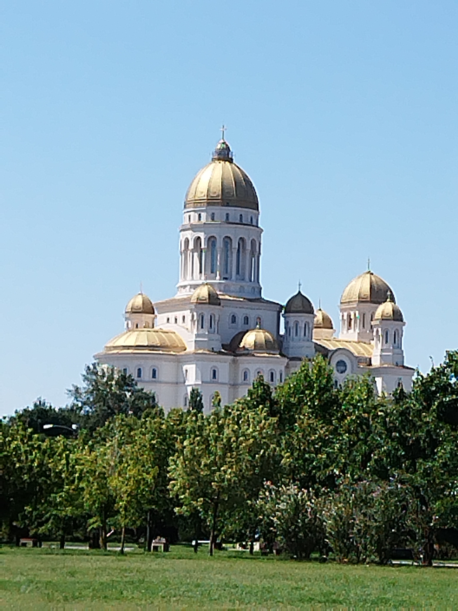 The image depicts the Salvation Cathedral, a large Orthodox Christian cathedral located in Bucharest, Romania. The cathedral is a dominant structure with multiple golden domes and white architectural elements, set against a clear blue sky. In the foreground, a green grassy lawn is visible, with several trees and some park benches scattered throughout the area. The scene appears to be a sunny day, with no people or specific actions prominently featured.