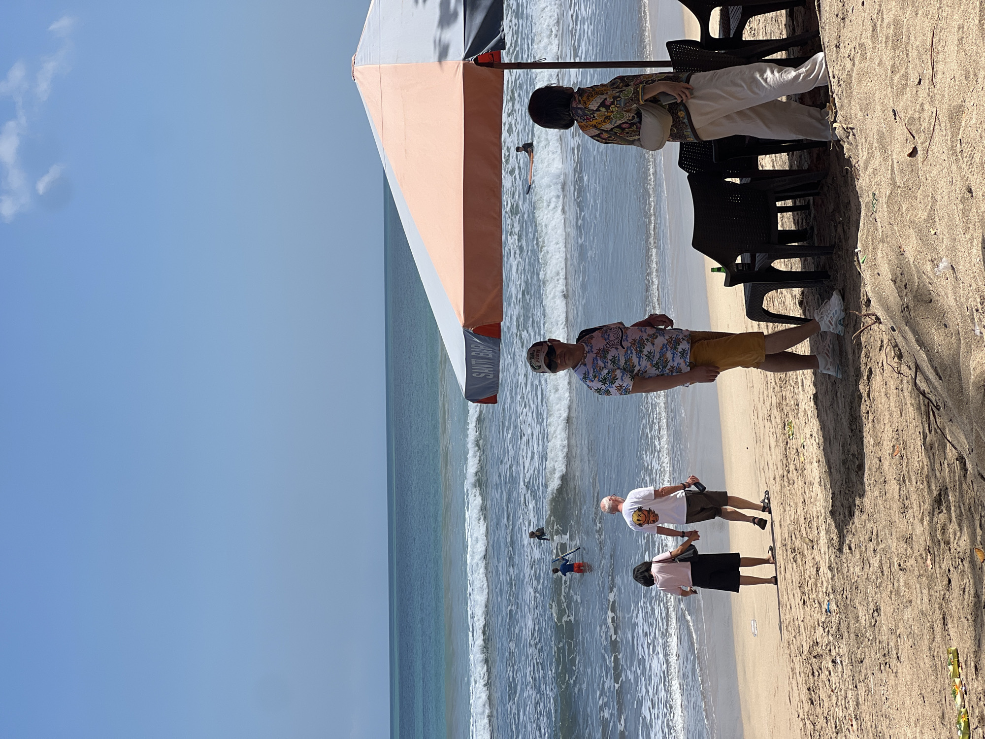 A bright, sunny day is captured on Kuta Beach, Indonesia, with a clear blue sky above a calm ocean. The foreground is a sandy beach with some scattered debris. Several people are present: a couple holding hands walks along the shoreline; a man in a tropical shirt and shorts stands near a beach umbrella, looking to his left; a woman in a colorful patterned shirt and light-colored pants stands next to him, facing away from the camera. Behind them, in the shallow water, a person in red shorts holds a surfboard, and another individual in dark clothing is further out in the ocean, possibly surfing. In the distance, the turquoise ocean meets the horizon under the expansive sky. A beach umbrella, orange and white, is set up on the sand, providing shade over a table and chairs, where a green bottle is visible.
