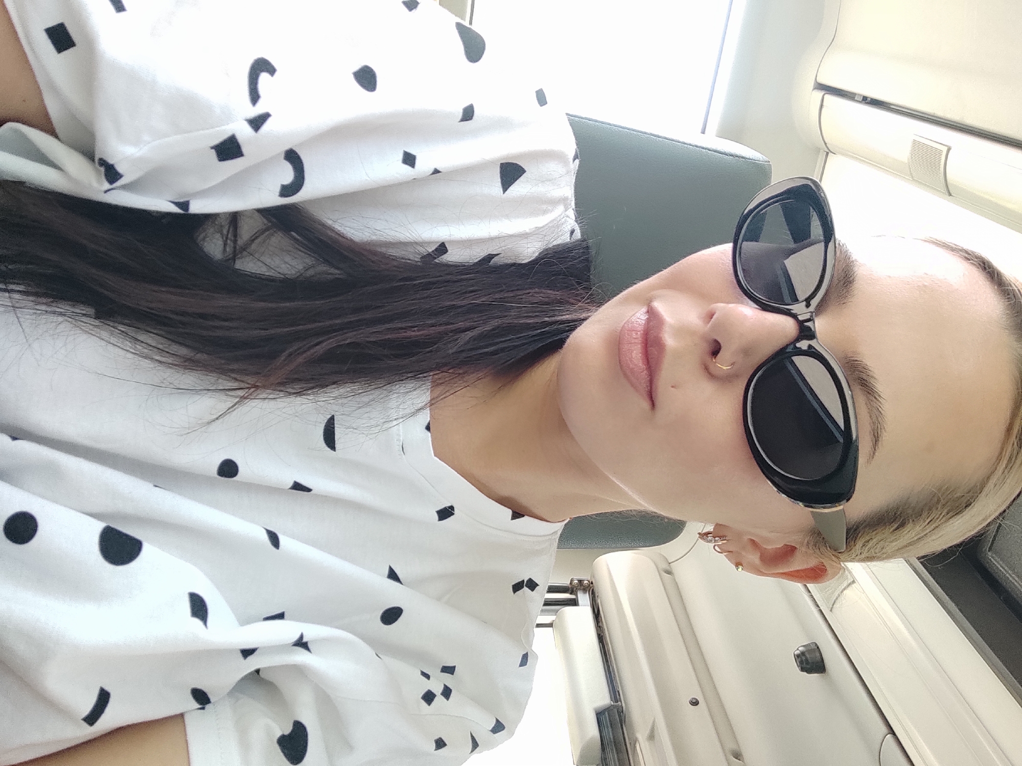 A selfie of a fair-skinned person with dark hair and blonde roots wearing sunglasses and a white shirt with black geometric patterns, taken indoors in what appears to be a train car, based on the interior paneling. The location in Chaikovsky, Russia, cannot be determined from the image.