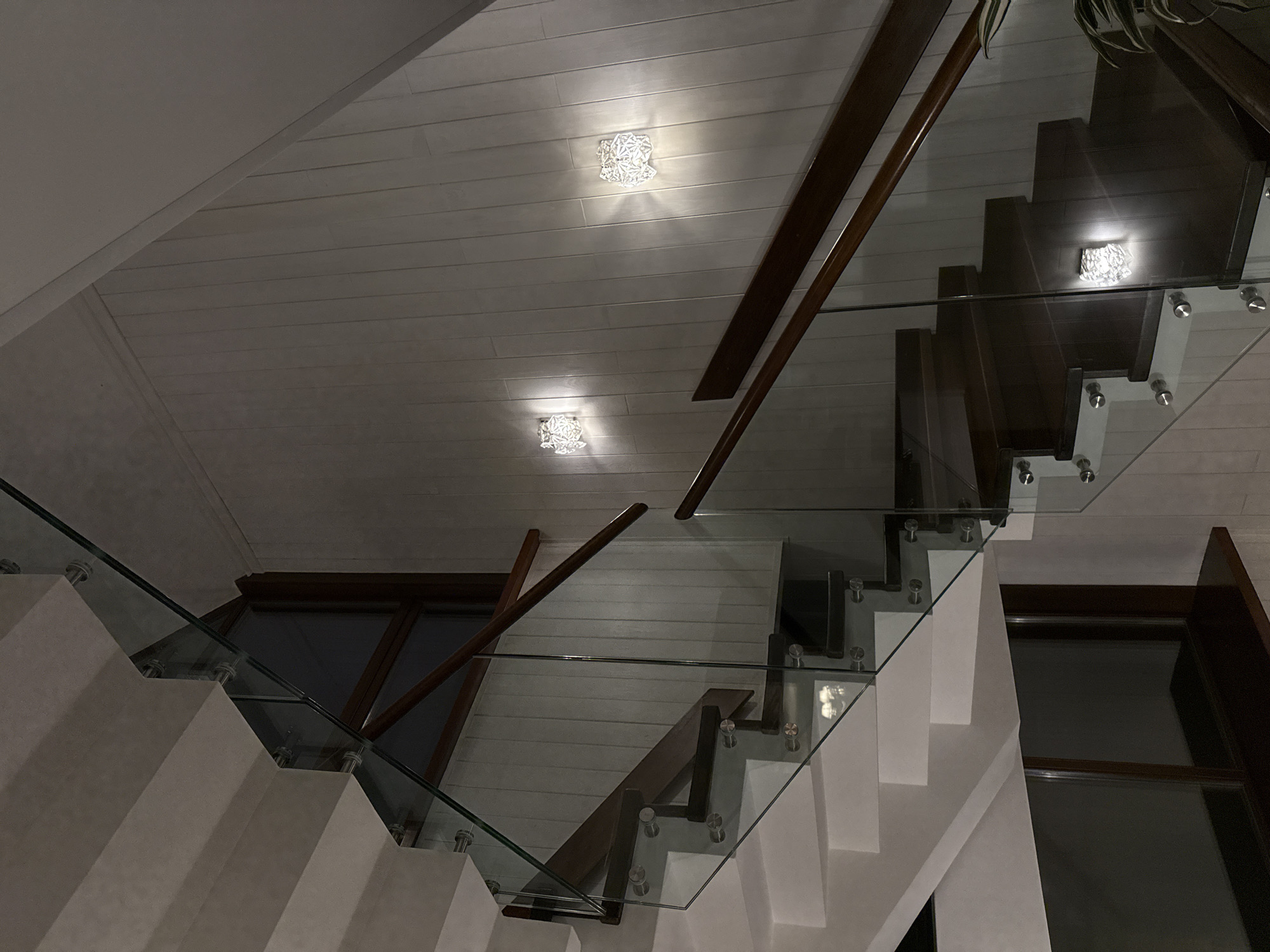 The image shows a modern staircase with dark wood steps and a glass railing. The staircase is illuminated by two decorative wall sconces, one on each side of the wall paneling. The wall is covered with vertical wood planks. The scene appears to be in a residential interior, possibly a home in Urlu-Aspak, Russia, although the location cannot be definitively confirmed from the image alone. There are no people visible in the image.