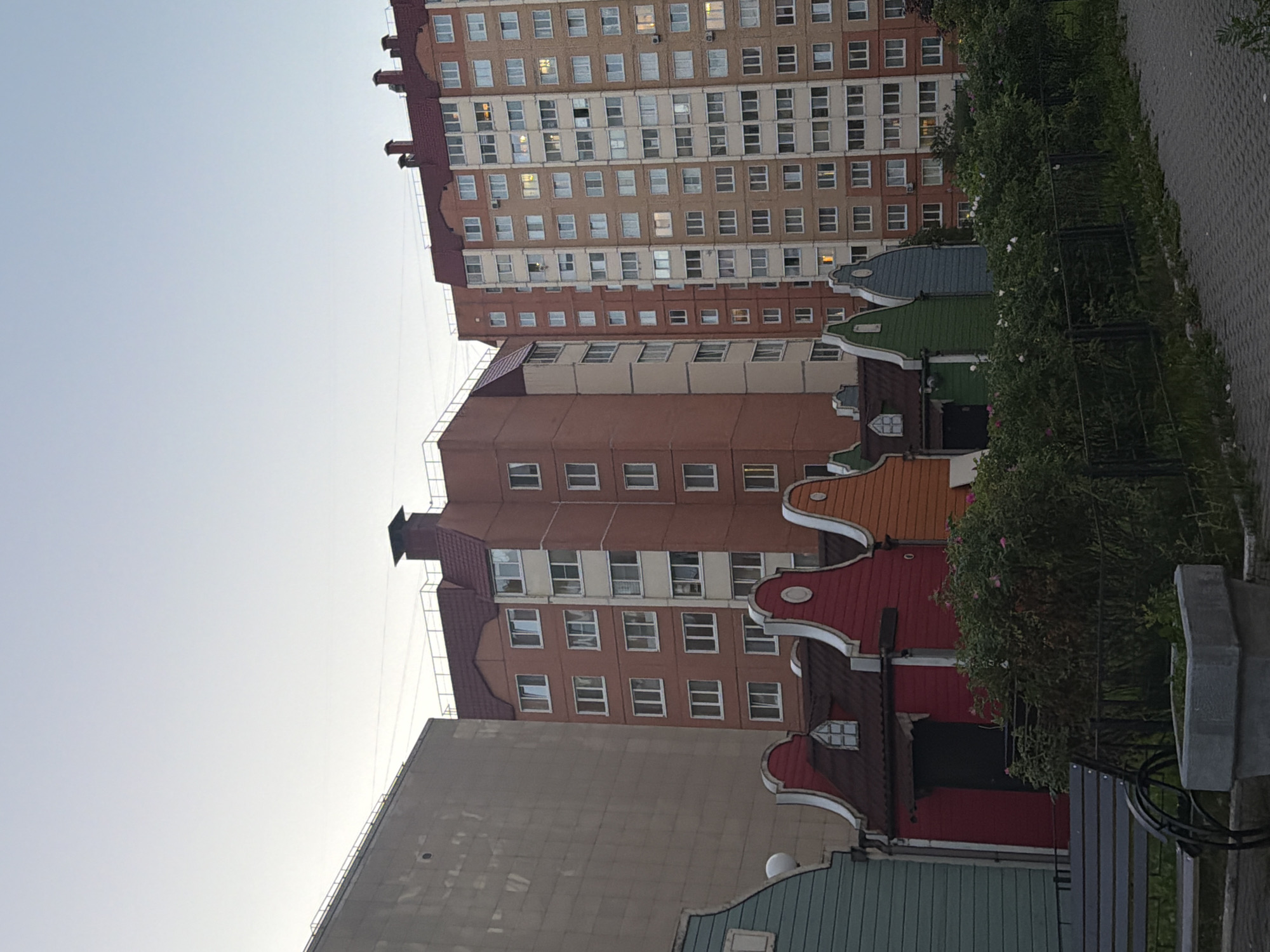 A Dutch-style row of small, colorful houses with stepped gable roofs is situated in front of a modern apartment building in a city. The houses are painted in vibrant colors like green, blue, red, and orange, and each has a distinct pitched roof with white trim. A dark doorway is visible on the red house, and a small window with a diamond-shaped pane sits above it. Behind these houses, a tall, multi-story apartment building with numerous windows dominates the background. The apartment building is a light brown color and features many balconies. To the left of the row of small houses, a large, beige building with a textured facade is partially visible. In the foreground, a dark metal bench is placed near a gray concrete planter, and a black metal fence runs along the edge of a paved walkway. Lush green bushes with pink and white flowers are growing in front of the fence. The sky above is a pale blue, suggesting it's either early morning or late evening.
