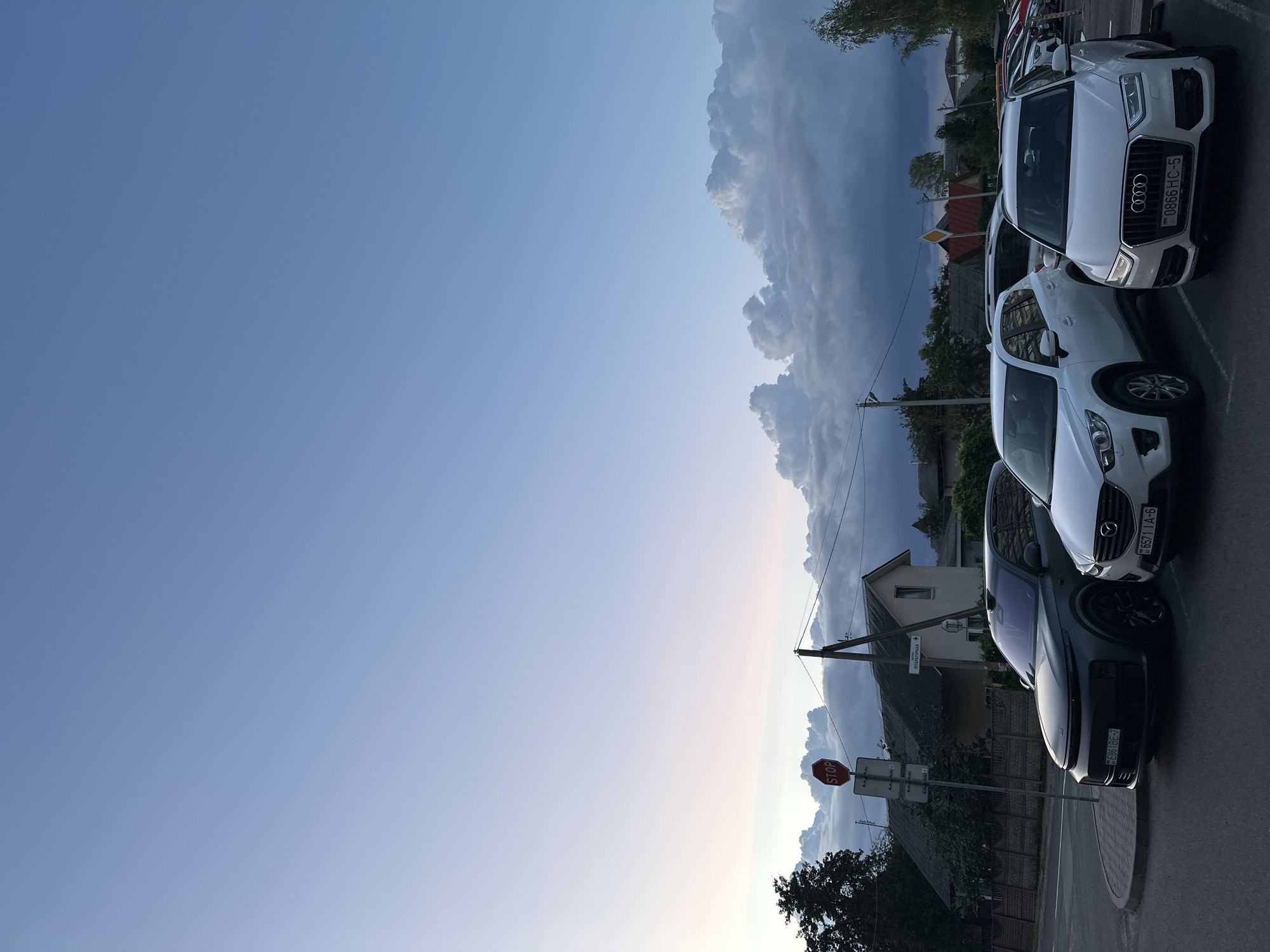 In Minsk, Belarus, at dusk, a parking lot scene unfolds under a vast, pale blue sky transitioning to a soft gradient of pink and lavender near the horizon. A band of dramatic, white cumulus clouds with dark grey undersides stretches across the sky, suggesting a recent or approaching storm. The foreground is occupied by several parked cars. Closest to the viewer is a white Audi Q3, its front grille featuring the iconic four rings logo and a license plate reading "0866 HC-5". To its left is a white Mazda CX-5 with a license plate "6571 IA-6". Further to the left, a dark grey SUV, possibly a Lynk & Co, is parked, its license plate partially visible as "BE861BE-7". Behind these vehicles, more cars are visible, some with their lights on. A red octagonal stop sign is prominently displayed on a metal pole to the left of the dark grey SUV, accompanied by a street sign. In the background, residential houses with dark roofs and trees are visible, their details softened by the twilight. Power lines crisscross the scene, connecting to utility poles. The overall atmosphere is calm, despite the dramatic cloud formations.