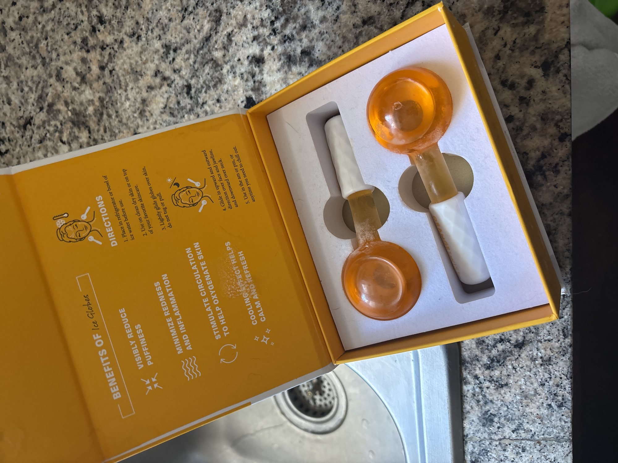 An overhead shot captures an open, rectangular yellow box containing two facial "Ice Globes" positioned on a speckled gray kitchen or bathroom countertop, with a stainless steel sink partially visible in the bottom left. The box features white molded inserts that cradle the two globes. Each globe has an orange, spherical top, a clear, light-orange stem, and a white, textured handle.
The inside lid of the box provides text about the product. On the left side, under the heading "BENEFITS OF Ice Globes," points include: "VISIBLY REDUCE PUFFINESS," "MINIMIZE REDNESS AND INFLAMMATION," "STIMULATE CIRCULATION TO HELP OXYGENATE SKIN," and "COOLING EFFECT HELPS CALM AND REFRESH." The right side of the lid, labeled "DIRECTIONS," outlines usage steps: "1. Place in refrigerator or bowl of ice water before use. 2. Use on clean dry skin or on top of your favorite skincare. 3. Lightly glide globes over skin, do not tug or pull. 4. Glide in upward and outward direction over face and jawline, and downward over neck. 5. Use in the am or pm, or anytime you need to chill out." The overall scene suggests a product ready for use in a personal care routine.