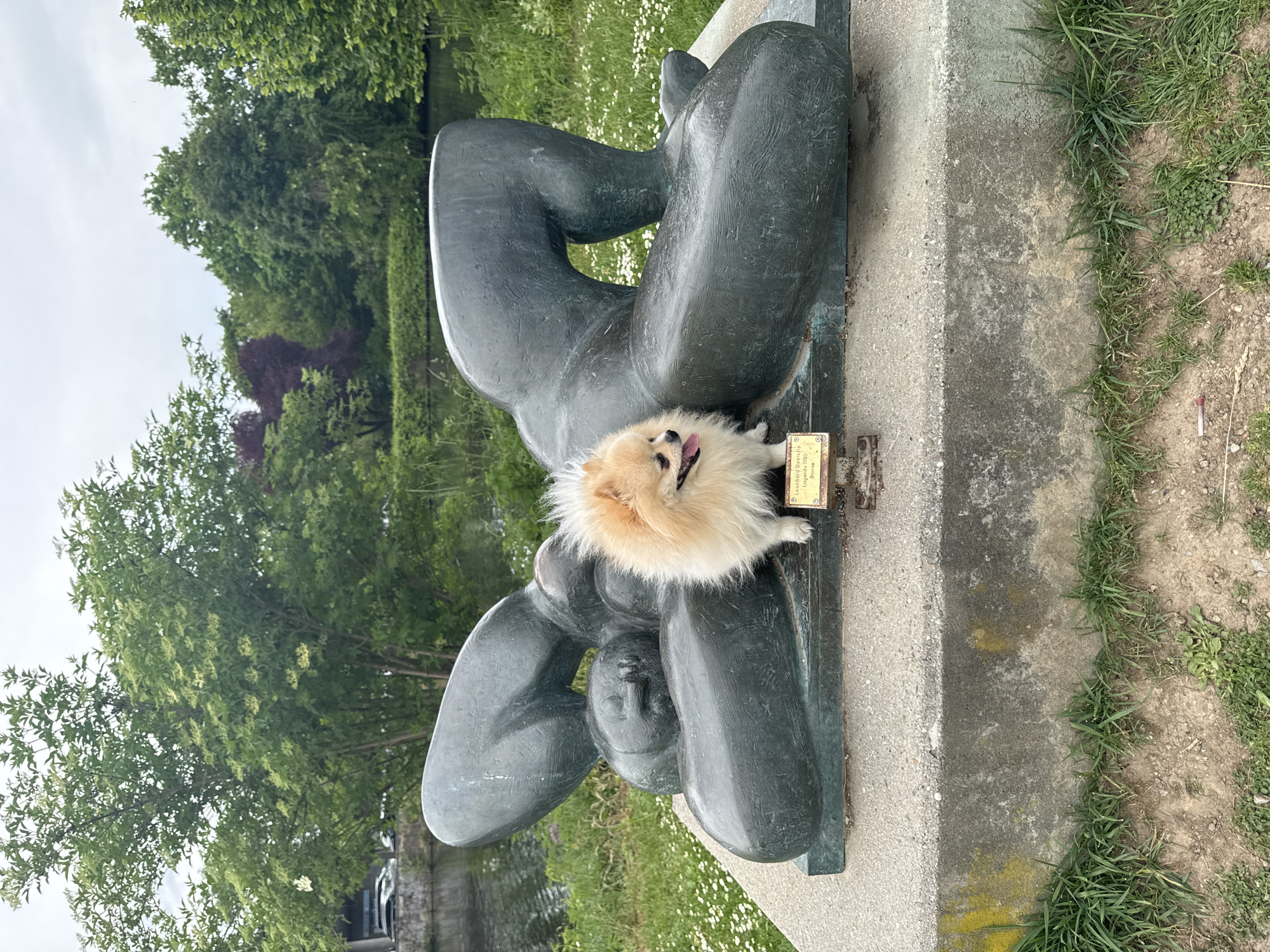 A Pomeranian dog is resting on a sculpture of a reclining female figure. The sculpture, made of dark metal, is mounted on a stone plinth. The dog is light brown with fluffy fur and its tongue is sticking out. A small metal plaque is affixed to the base of the sculpture. The sculpture is located in a grassy area with trees in the background. The location is Bietigheim-Bissingen, Germany.
