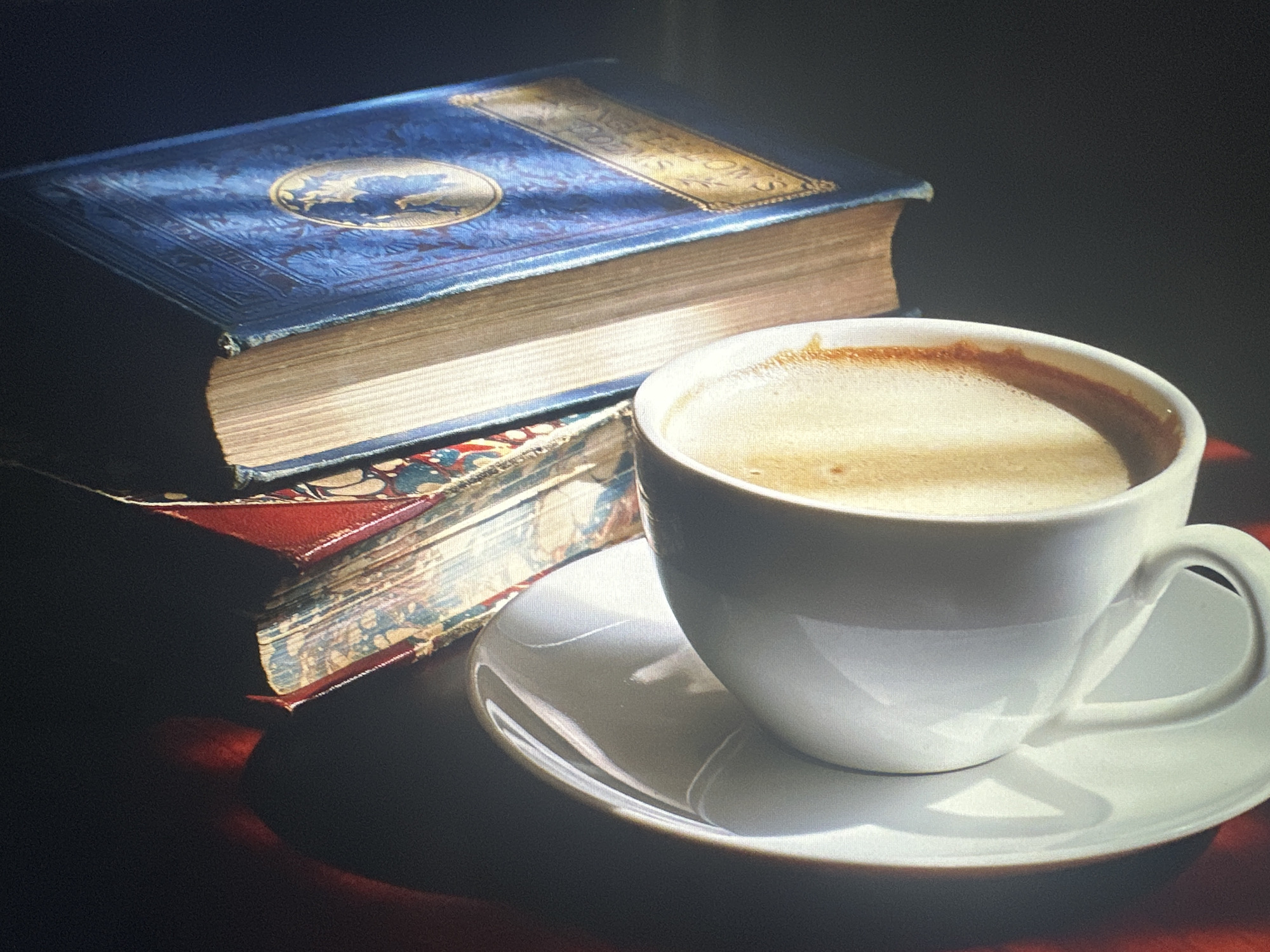 The image depicts a still life composition featuring two antique books and a white cup of coffee on a saucer. The books are stacked, with the top book being larger and bound in blue with decorative gold embossing. The lower book is partially visible, appearing to have a red cover and ornate marbled endpapers. The coffee cup is white ceramic and filled with a foamy beverage, casting a shadow on the white saucer. The scene is lit by natural light, creating dramatic shadows and highlights, suggesting a window or direct light source from the left. The background is dark and out of focus, emphasizing the main subjects. The overall mood is serene and contemplative. There is no discernible location in the city of Bryansk, Russia, present in this image; it is a generic still life arrangement.