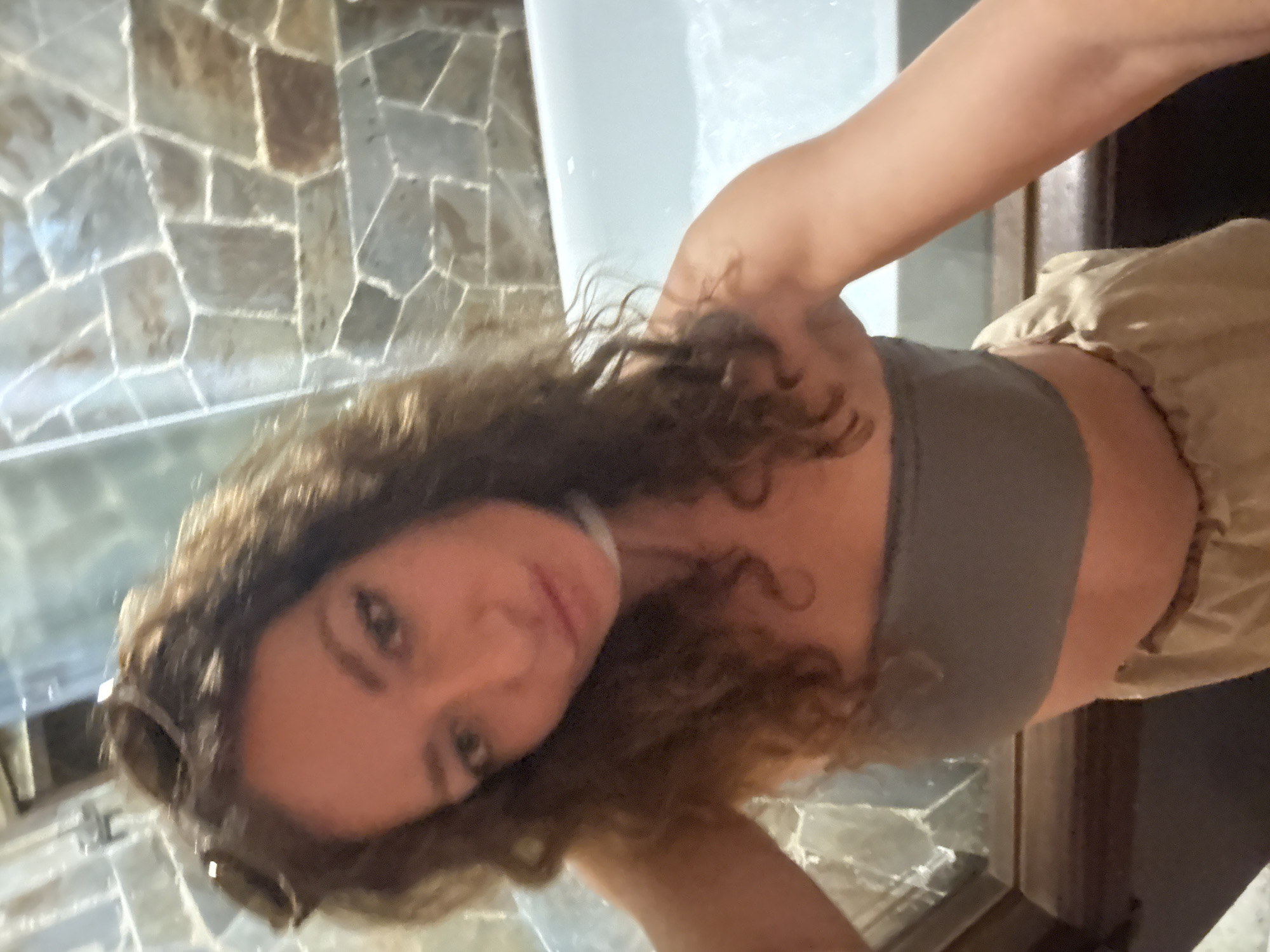 A female subject is depicted in a selfie taken indoors in Mù Cang Chải, Vietnam, likely within a bathroom. The woman has long, dark, curly hair with sunglasses perched on her head. She is looking directly at the camera with a slight pout. She wears a grey strapless crop top, light-colored loose pants or skirt, and a white choker. Her arms are visible, with her right arm (viewer's left) raised to hold the camera. The background features a wall constructed from irregular, multi-toned stone tiles with white grout lines. To the subject's right, a white bathtub is partially visible, containing water and foam. A dark brown ledge or counter runs below the bathtub and extends across the lower left portion of the frame.