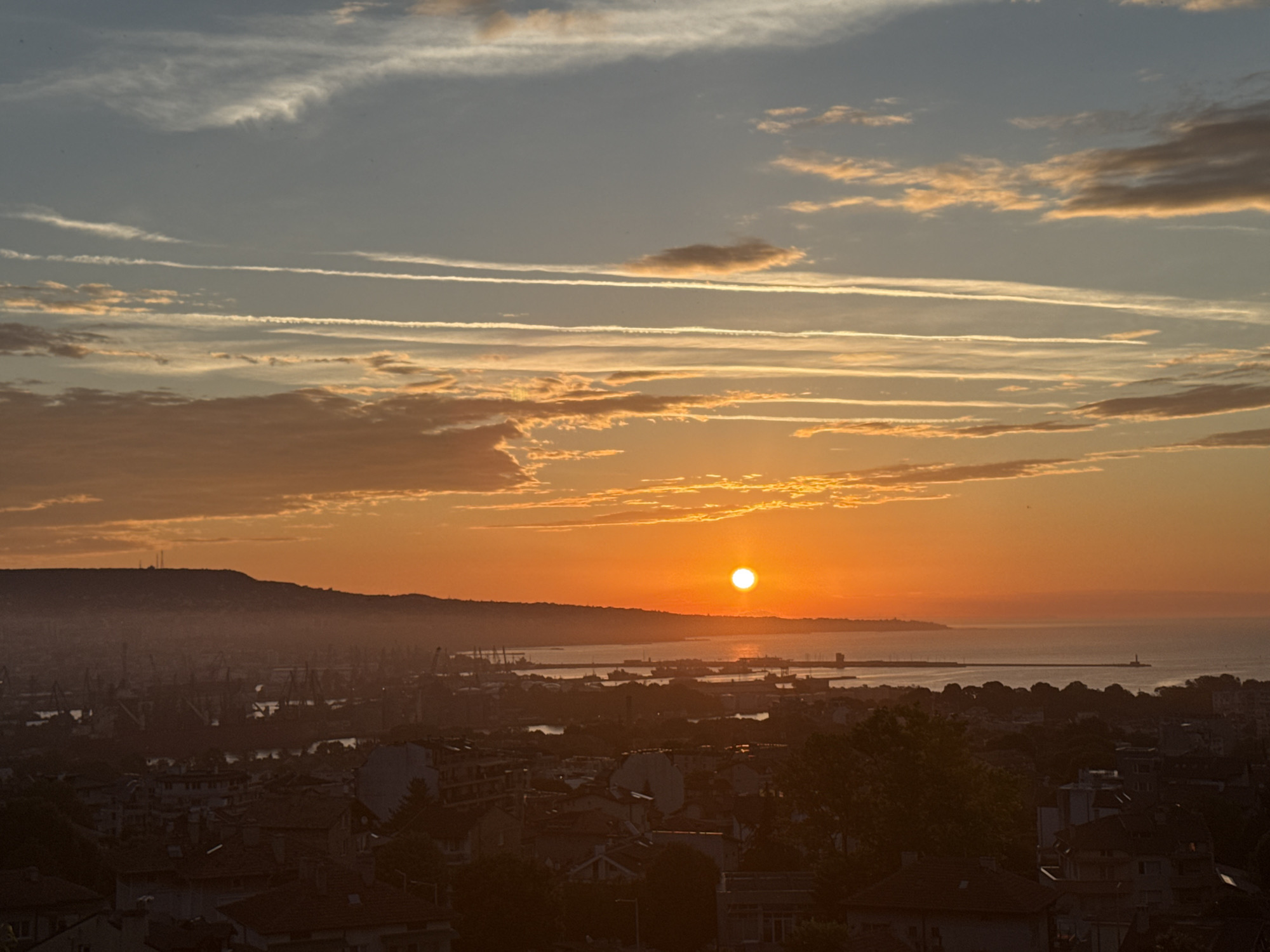 The image captures a vibrant sunset over a coastal city, likely Unknown, Unknown. The sky is a spectacle of warm hues, with the sun a brilliant orb near the horizon, casting a golden glow across the scene. Wispy clouds, some tinged with orange and pink, stretch across the sky, interspersed with streaks of contrails.
Below the fiery sky lies a sprawling cityscape, densely packed with buildings. The architecture appears to be a mix of older structures with tiled roofs and more modern apartment buildings. Trees are scattered throughout the urban landscape, adding pockets of green amidst the rooftops.
In the mid-ground, a large port is visible, teeming with ships and cranes, suggesting industrial activity. The water of the harbor reflects the warm colors of the sunset. The background is dominated by a hazy, mountainous silhouette, with the mist softening its edges and adding depth to the scene. The overall impression is one of a dynamic urban environment bathed in the dramatic light of a setting sun.