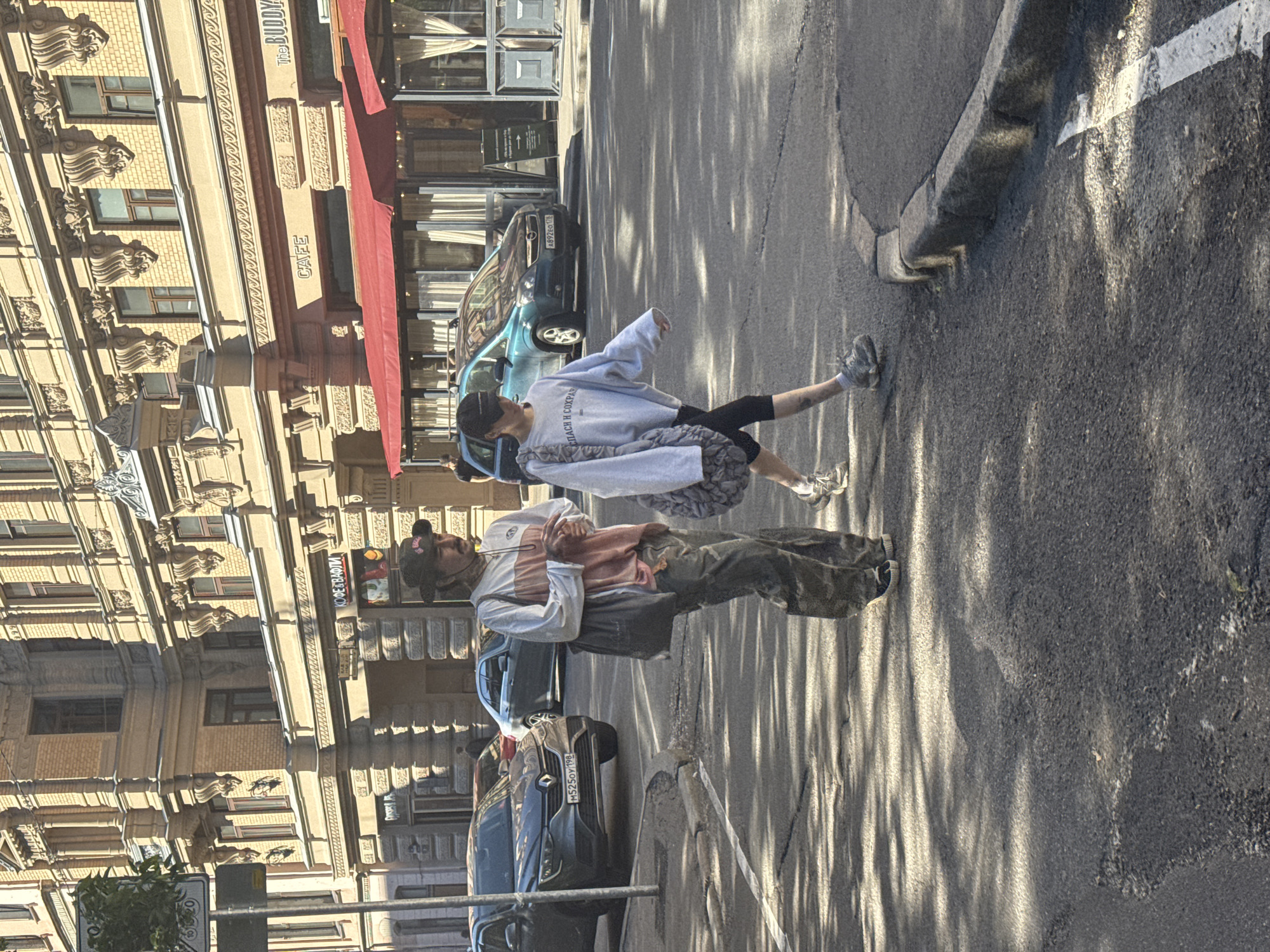The image depicts a street scene in Saint Petersburg, Russia. Two individuals are the primary subjects. One person, wearing a grey sweatshirt with Cyrillic text, black leggings, and sneakers, is captured mid-stride, walking across the street. They are carrying a bag with a patterned strap. The second person, dressed in a striped long-sleeved shirt, pants, and a baseball cap, stands looking down and holding an object.
The setting is an urban environment with a classic European architectural style evident in the building facade on the left. This building features ornate detailing, multiple windows, and a red awning extending from one of the establishments. The street is paved with asphalt and marked with a white line. Shadows cast by trees or structures create a dappled pattern on the road, suggesting it is daytime. Two cars are visible on the street, one a teal-colored vehicle with a license plate, and another darker car parked further down.
Visible text includes "THE BUDDY" and "CAFE" on the building, and Cyrillic text on the sweatshirt of the person walking. Another visible license plate reads "M525OY 198". The overall mood appears casual and ordinary, a snapshot of everyday life in the city. The lighting indicates it is a sunny day.