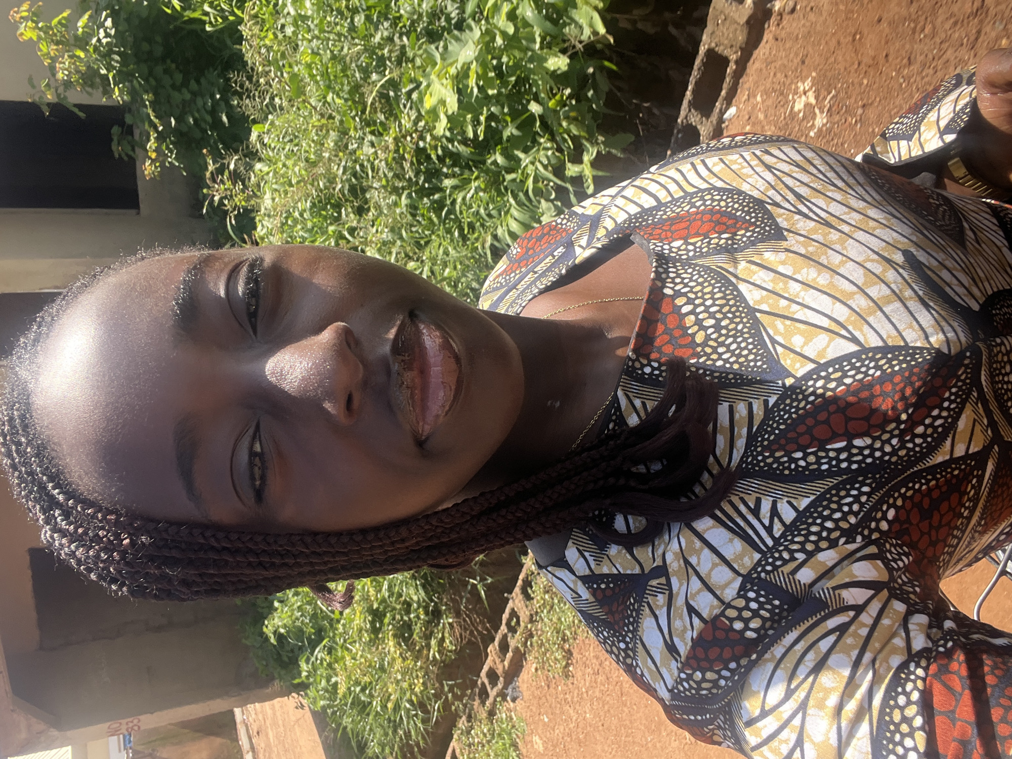 An outdoor portrait in Abuja, Nigeria, features a dark-skinned woman with braided hair, facing forward and looking directly at the camera with a slight smile. She is wearing a vibrant, multi-colored patterned garment, consistent with Ankara fabric, featuring white, yellow, black, and reddish-brown hues in abstract and circular motifs. A delicate gold-colored chain necklace is visible around her neck, and a gold-colored watch adorns her left wrist.
The scene is brightly illuminated by natural sunlight, casting subtle shadows. To the viewer's left, a portion of an unpainted or unfinished concrete building is visible, with a partially obscured white sign displaying "NO. 33" in black text. The ground in the foreground and to the left is composed of reddish-brown earth, indicative of laterite soil. To the viewer's right and in the background, dense green vegetation, appearing as overgrown bushes and plants, fills the frame. Behind the vegetation, another indistinct dark structure, possibly part of a building, is partially visible. Concrete blocks are scattered on the reddish-brown ground to the left and rear of the woman.