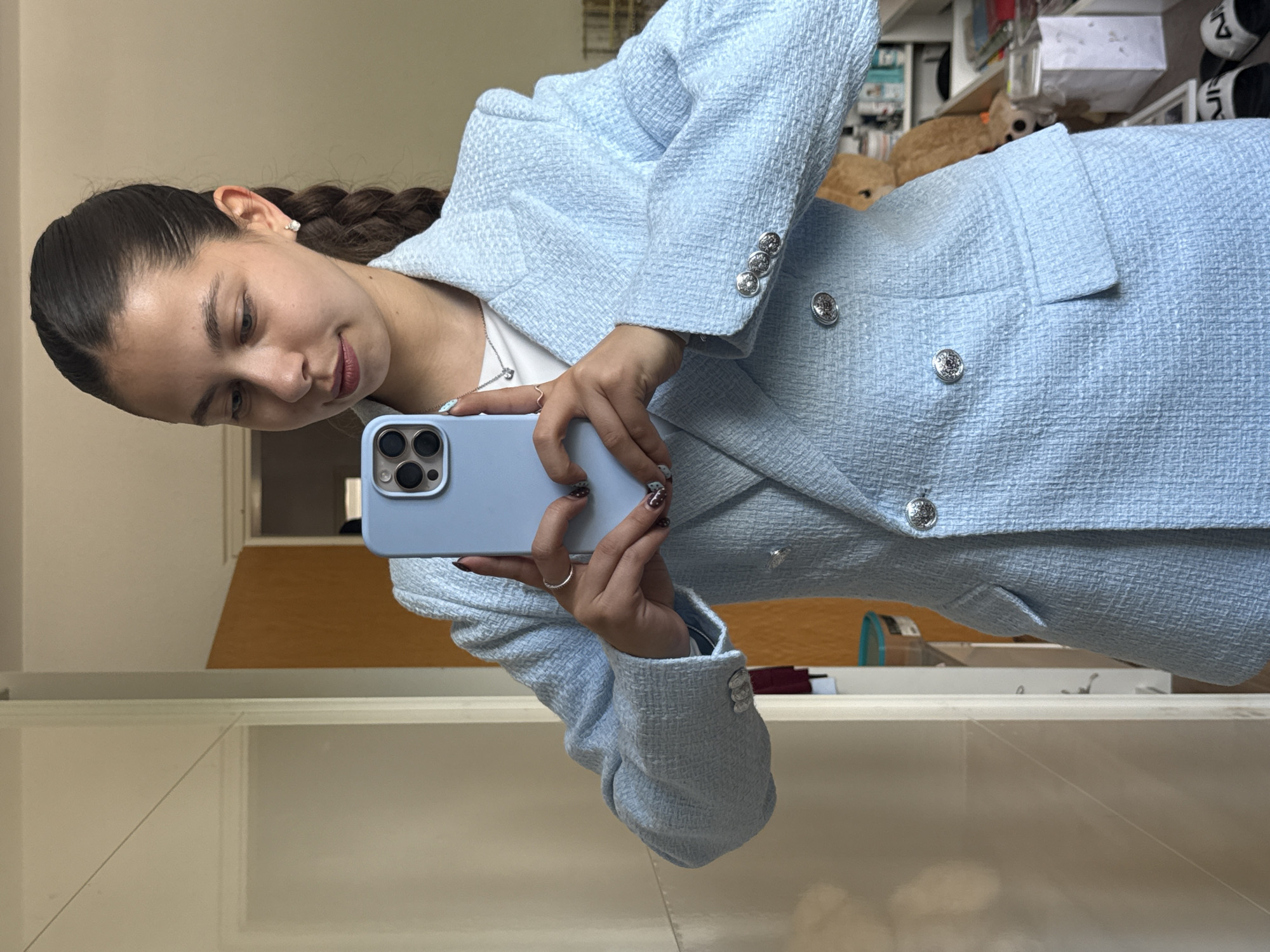 A young woman is taking a selfie in a mirror. She is wearing a light blue tweed blazer and skirt with silver buttons. Her dark hair is pulled back in a braid. She holds a light blue iPhone in her hands. The background appears to be a room with shelves and various objects, but is blurry. The location is Bruchköbel, Germany.