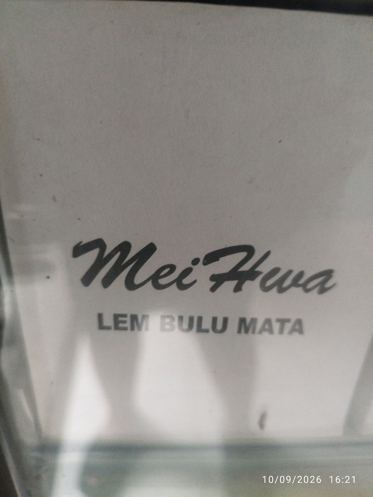 This is a close-up, slightly blurry photograph of a white surface with black text. The main text in a stylized script reads "Mei Hua". Below this, in block letters, it says "LEM BULU MATA". The bottom right corner of the image displays a date and time stamp: "10/09/2026 16:21". There is no discernible environmental context to suggest a specific location within the city of Mojokerto, Indonesia. The image is a static depiction of text on a surface.