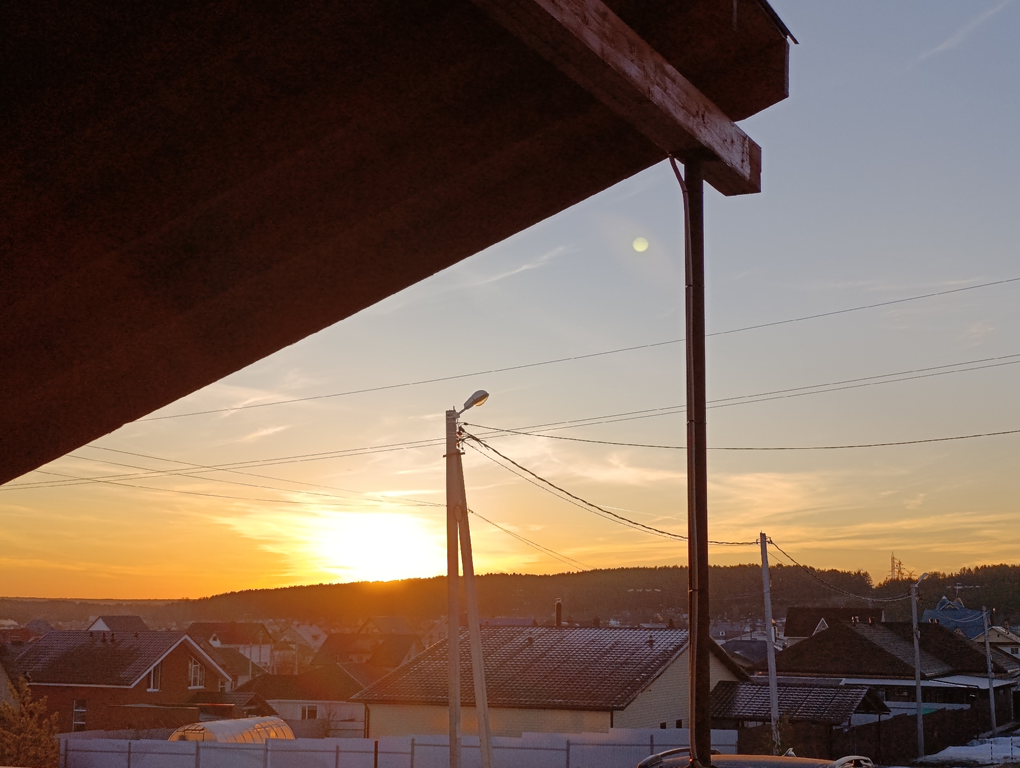 The image shows a sunset over a residential area, likely in the city of Chaikovsky, Russia. The sun is setting on the horizon behind a line of trees, casting a bright orange glow on the sky and clouds. Several houses are visible in the foreground, with their roofs partially covered in snow. Power lines stretch across the sky, and a street lamp is positioned on a pole. The image is partially framed by a dark brown wooden structure in the upper left corner.