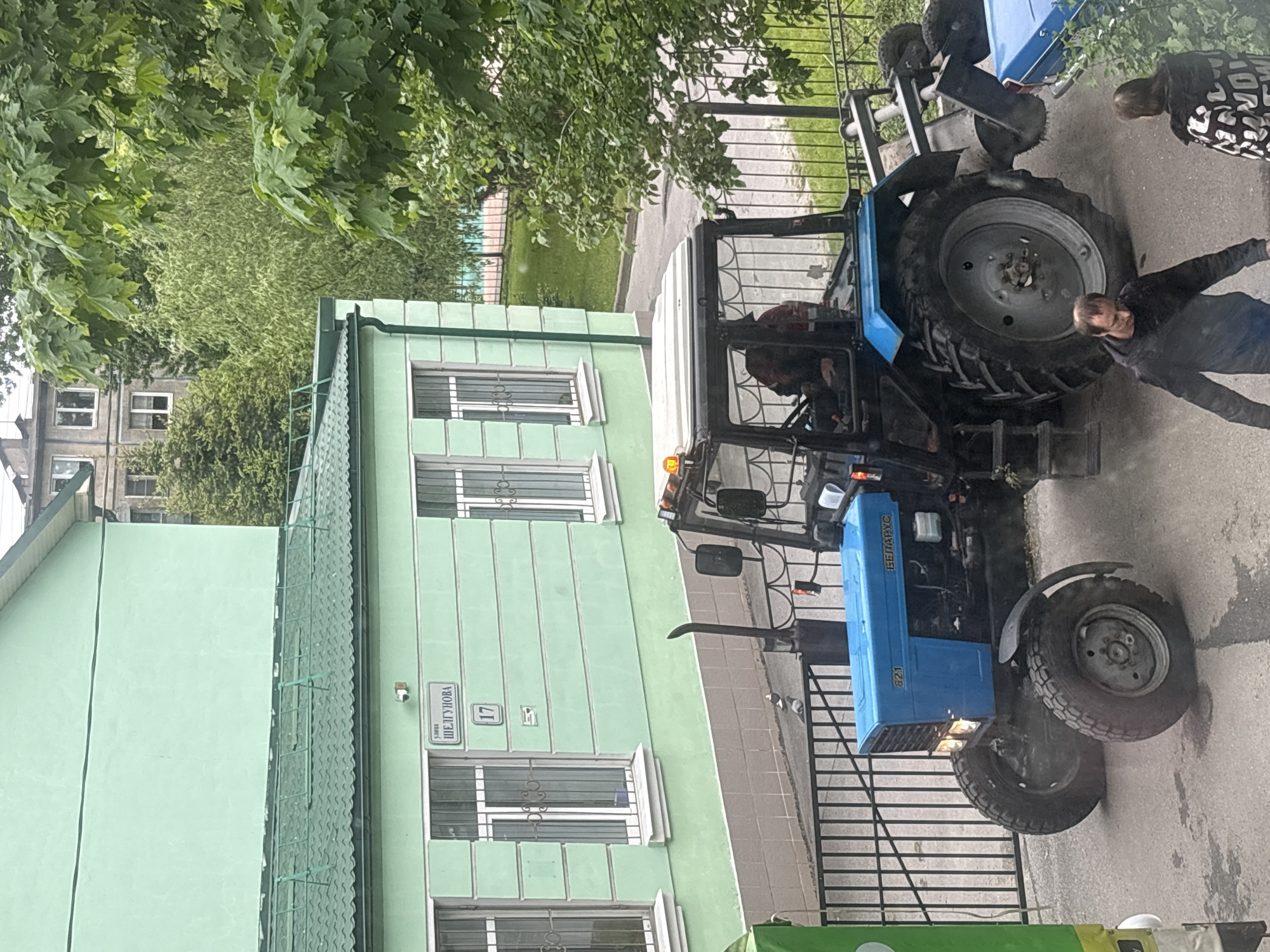The image depicts a blue Belarus 82.1 tractor parked on a sidewalk in front of a green building. The building, located at 17 Shelgunova Street in Saint Petersburg, Russia, features multiple windows with white frames. Two people are near the tractor, one standing and another leaning towards the large tractor tire. A tree is visible in the background. The tractor appears to be in an urban setting, beside a building and near a metal fence.