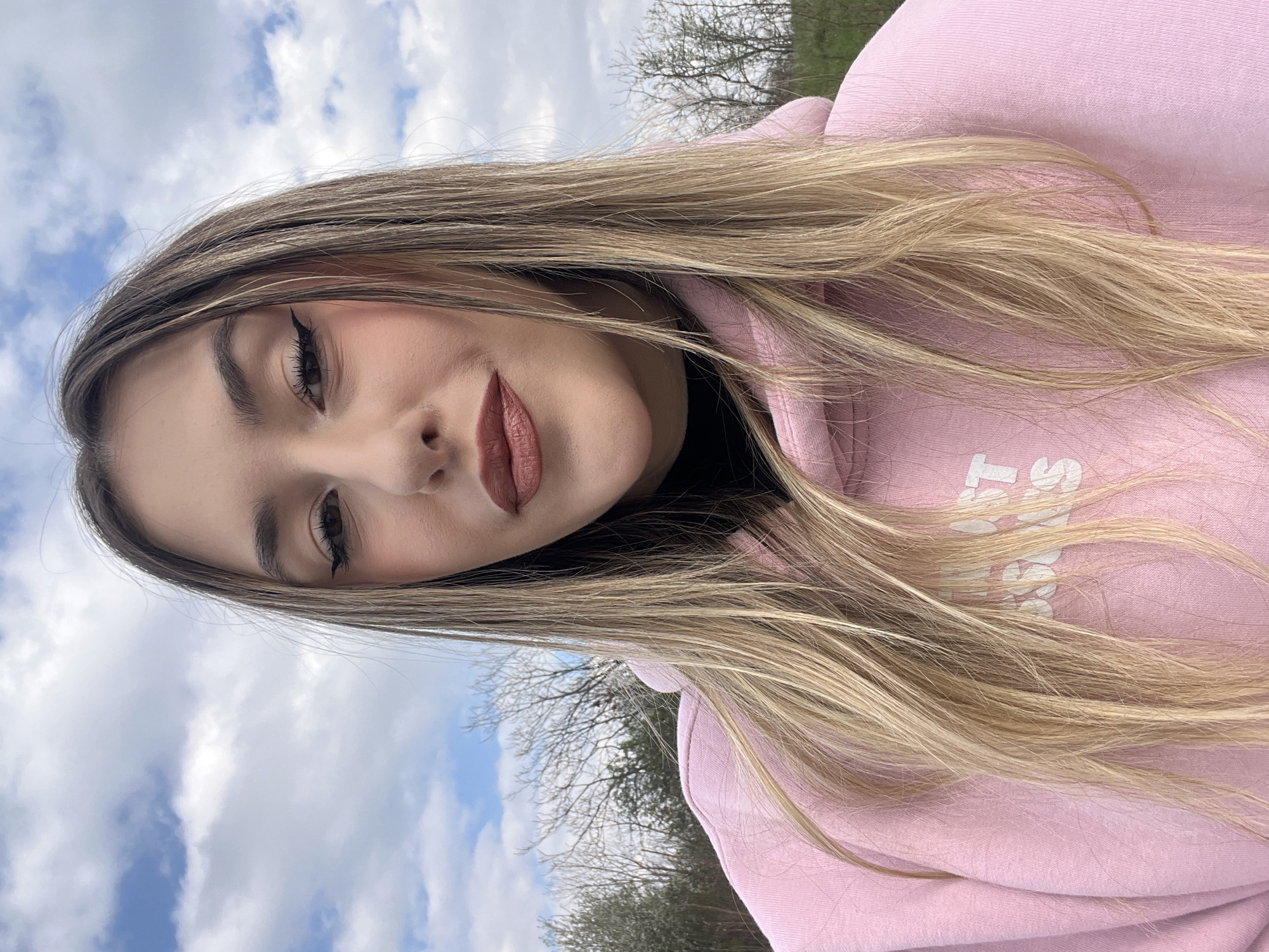 A young woman with light brown hair is wearing a pink hoodie. She is angled slightly to the left and looking down at the camera. The background shows a blue sky with clouds and trees with bare branches. The location is likely a park or green space in Sofia, Bulgaria, given the trees and sky visible in the background.