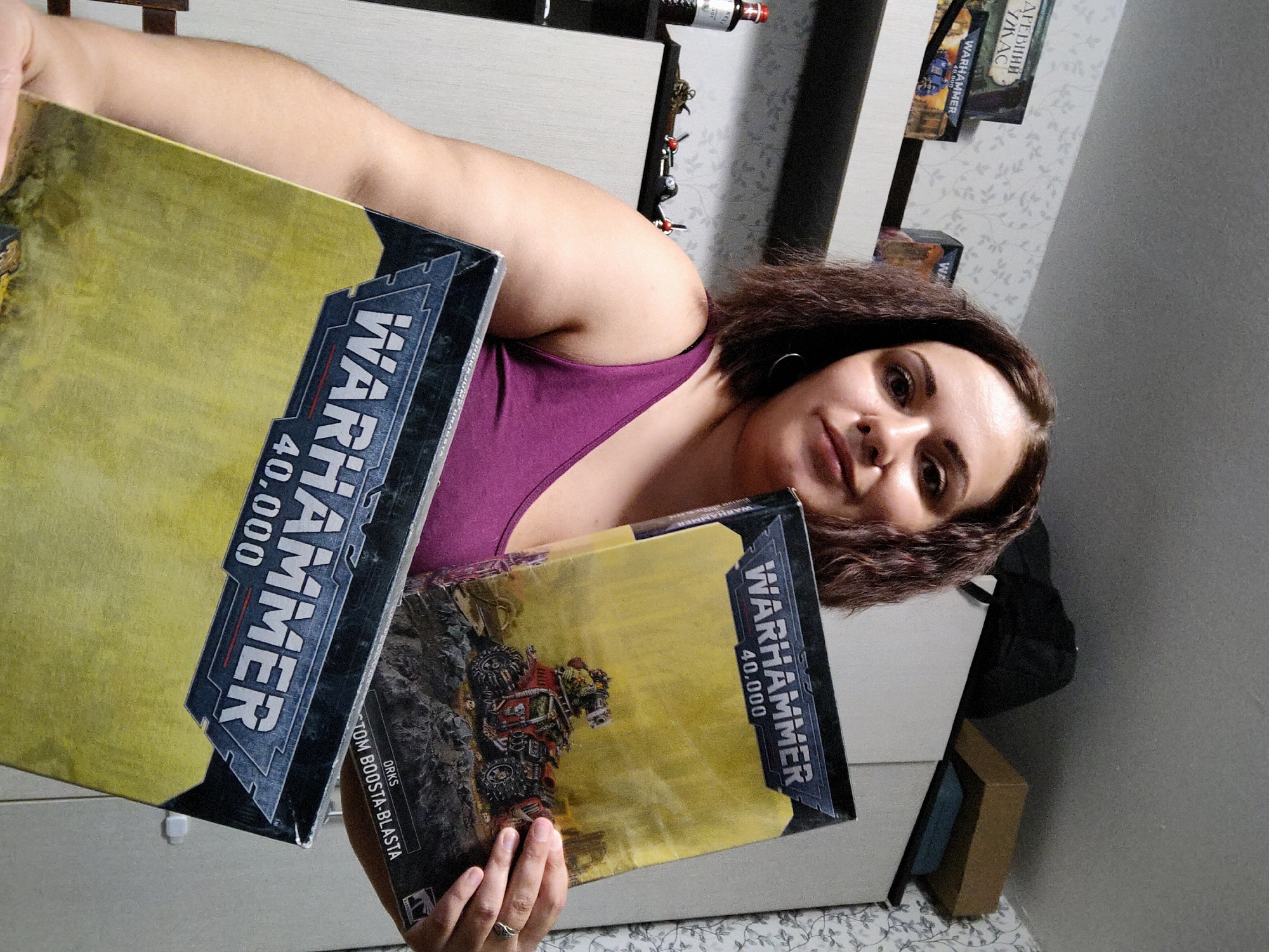 A person in a purple tank top holds two "Warhammer 40,000" game boxes. The person is indoors, possibly in a home. A cabinet or storage unit is behind the person, with miscellaneous items on top of it, including a bottle and other "Warhammer" boxes. The background wall has a floral pattern. There are no specific details that confirm the location is in Марковский, Russia.