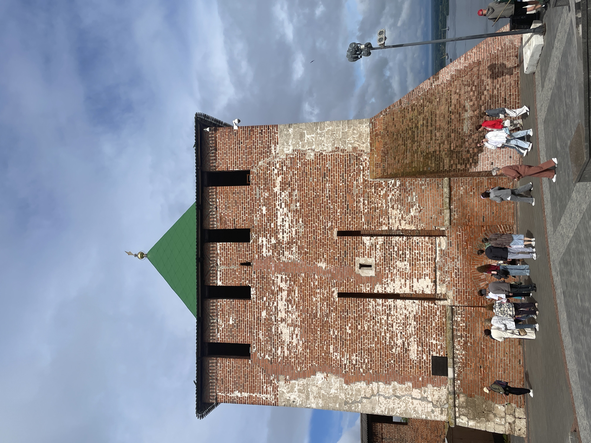 A brick tower with a green roof and a gold finial stands prominently under a cloudy sky. The tower's brickwork shows signs of wear, with patches of white plaster visible. Several dark, rectangular openings dot the upper section of the tower, serving as windows or battlements. A large, angled brick structure juts out from the right side of the tower. In the foreground, a paved area has a group of people walking and standing. To the right of the tower, a tall lamppost with lights is visible. In the distance, a body of water, likely a river, stretches towards the horizon. The scene is set in Nizhny Novgorod, Russia, specifically within the Nizhny Novgorod Kremlin, a historic fortress.