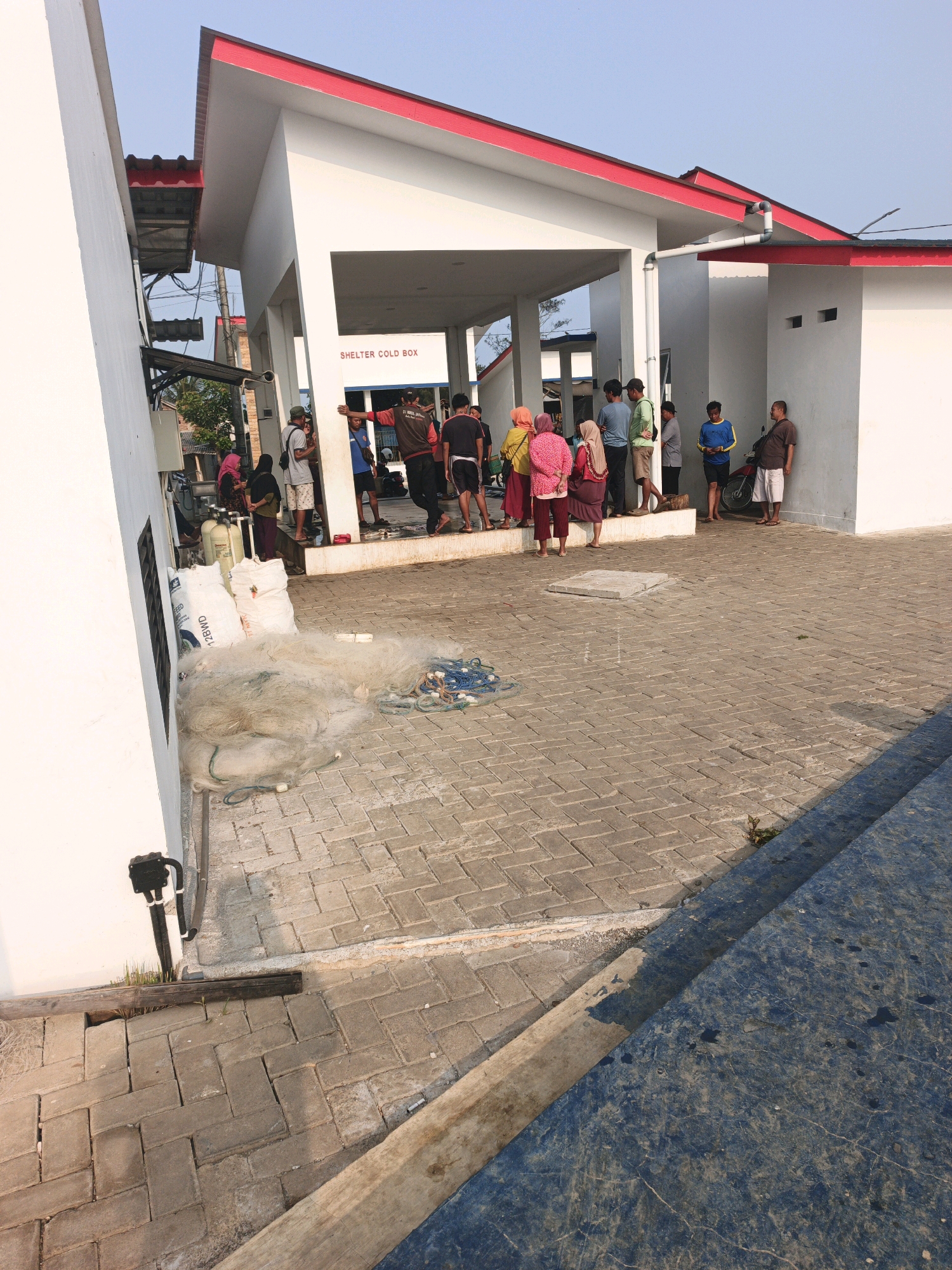 In an outdoor area paved with interlocking bricks, a group of people stand around a covered shelter with a red roof. The shelter has the words "SHELTER COLD BOX" written above an opening. To the left of the shelter, two white sacks and a pile of fishing nets are visible on the ground. Several individuals are standing near the shelter entrance, some appearing to be in conversation. A motorcycle is parked near the right side of the scene. The overall environment suggests a service area or community space, possibly related to fishing or goods storage. The time of day appears to be late afternoon, given the lighting and long shadows.