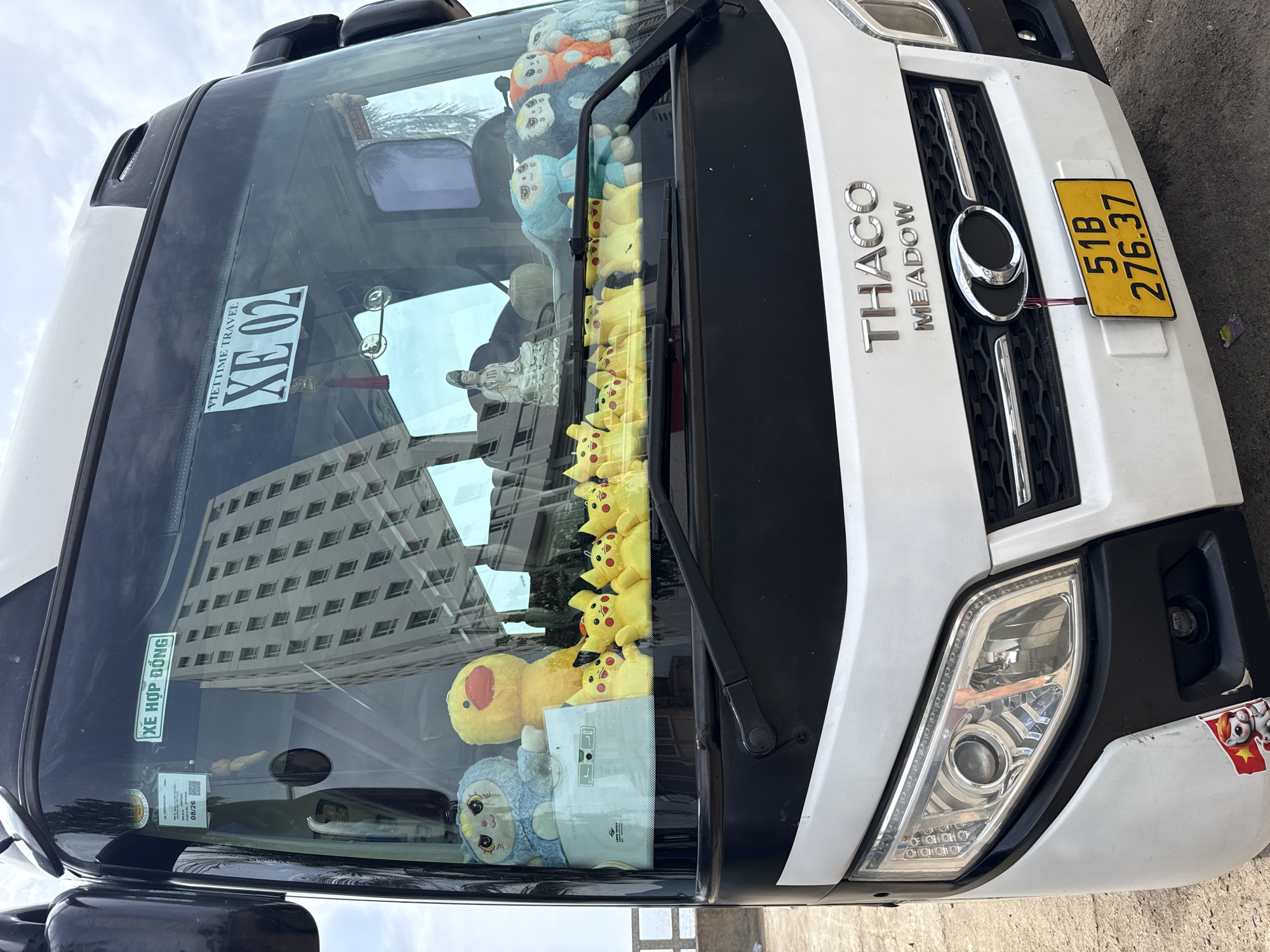 This is a front-view, eye-level, medium shot of a white and black THACO MEADOW bus parked outdoors. The bus's windshield is decorated with numerous yellow plush Pikachu toys, a large yellow duck toy, and a blue plush cat toy. A small white Buddha statue and a light green decorative object are also visible inside the windshield.
Above the windshield, a white rectangular sticker with Vietnamese text and the number "XE 02" is affixed. To the left, another sticker with Vietnamese text and a QR code is visible. A yellow license plate with the text "51B 276.37" is mounted on the lower front of the bus. The background shows a cloudy sky and a tall building reflected in the windshield. The bus appears to be parked on an asphalt surface with a concrete barrier on the left side. There is no indication of the specific location in Nha Trang, Vietnam, within the image itself.