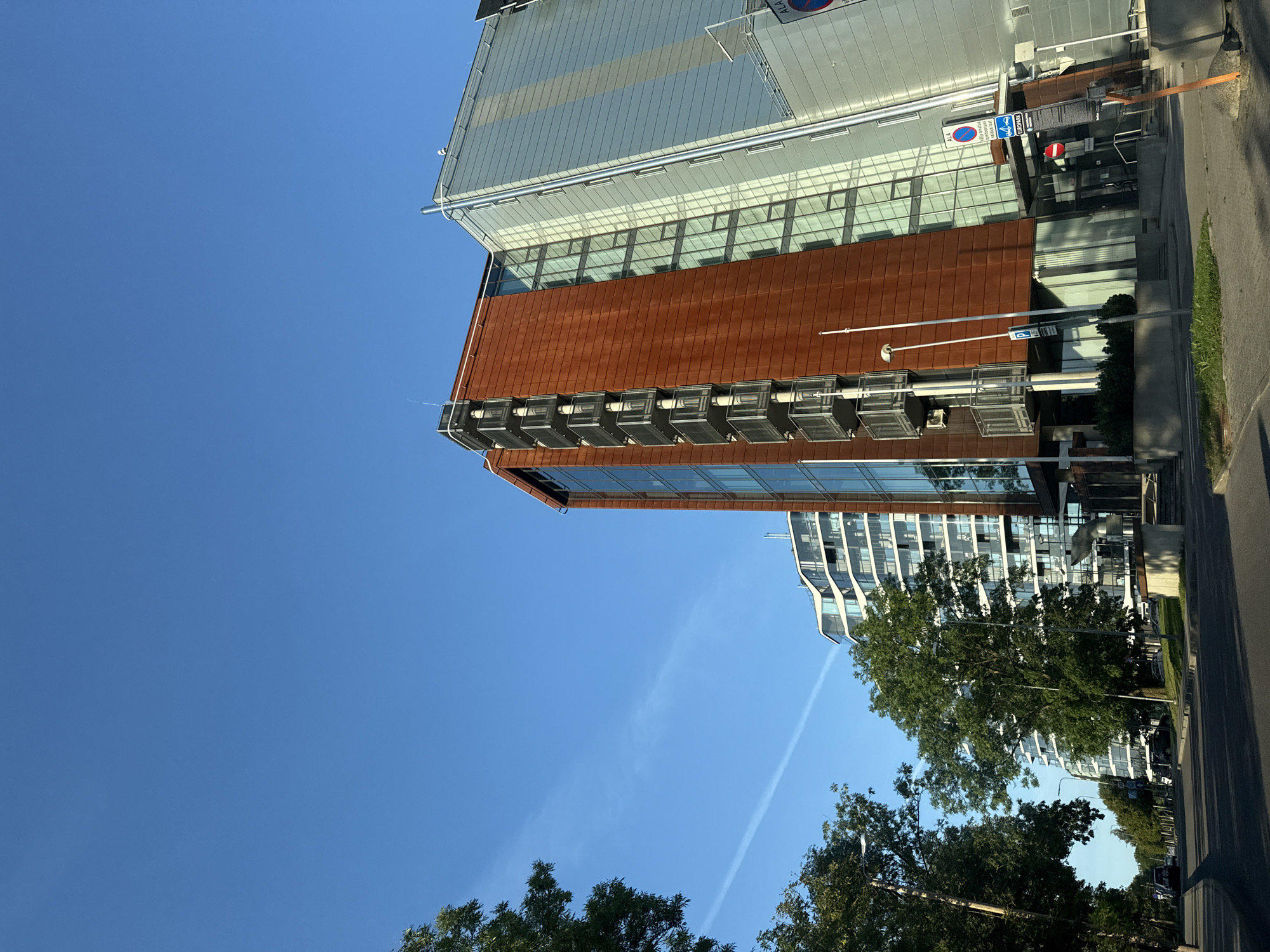 The image shows a modern residential building with a distinctive reddish-brown facade and glass elements, situated in a city environment. In the foreground, a street with cars and trees lines the side. To the left of the reddish-brown building, another contemporary residential building with a curved white facade is visible. The sky is clear blue with a faint contrail. The architecture suggests a relatively new development in a city known for its modern urban planning. Based on the provided context, this scene is located in Tallinn, Estonia, specifically in the Nauka Maja area, which is characterized by modern apartment buildings and a mix of residential and commercial spaces. The building with the reddish-brown facade is identifiable as a prominent structure in this area, featuring balconies with metal railings. The overall impression is of a well-maintained urban landscape with contemporary architectural design.