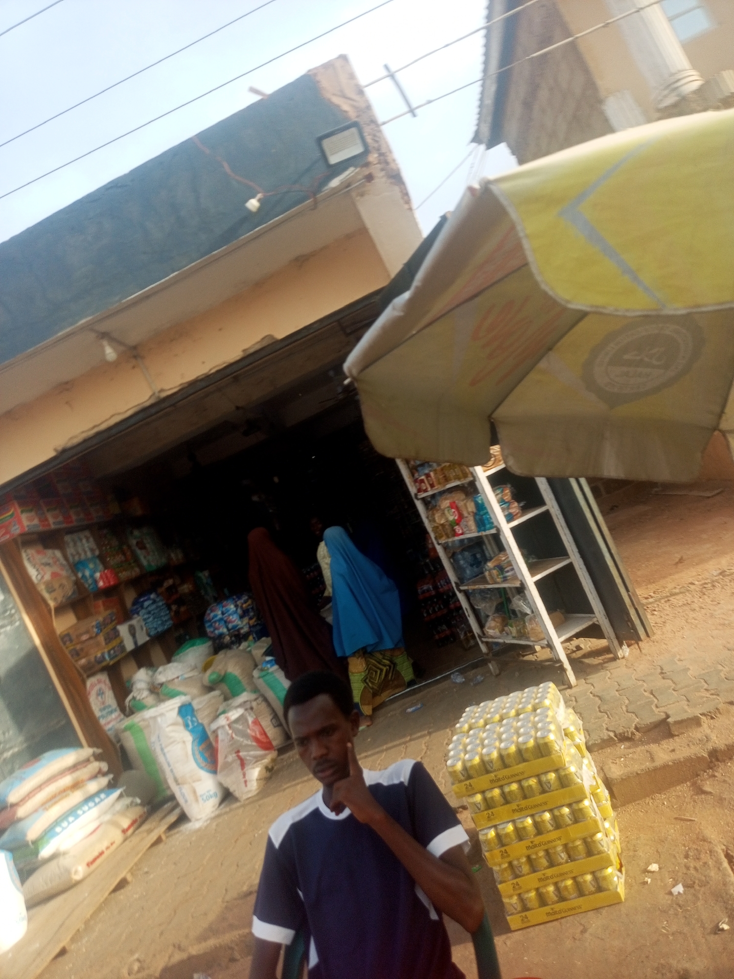 Here is a concise description of the image:
A medium shot captures a man seated in front of what appears to be a small convenience store in Unknown, Unknown. The store is open, with shelves visible inside stocked with various items. Two individuals in traditional attire are browsing the shelves inside the store. Bags of products, including a bag labeled "BUA SUGAR 50kg" are stacked in front of the store entrance. A stack of canned beverages stands to the right of the man. An umbrella with branding, partially obscures the top right of the frame, and is positioned in front of the shop. The scene is shot during the day, with natural lighting casting shadows on the ground and building.