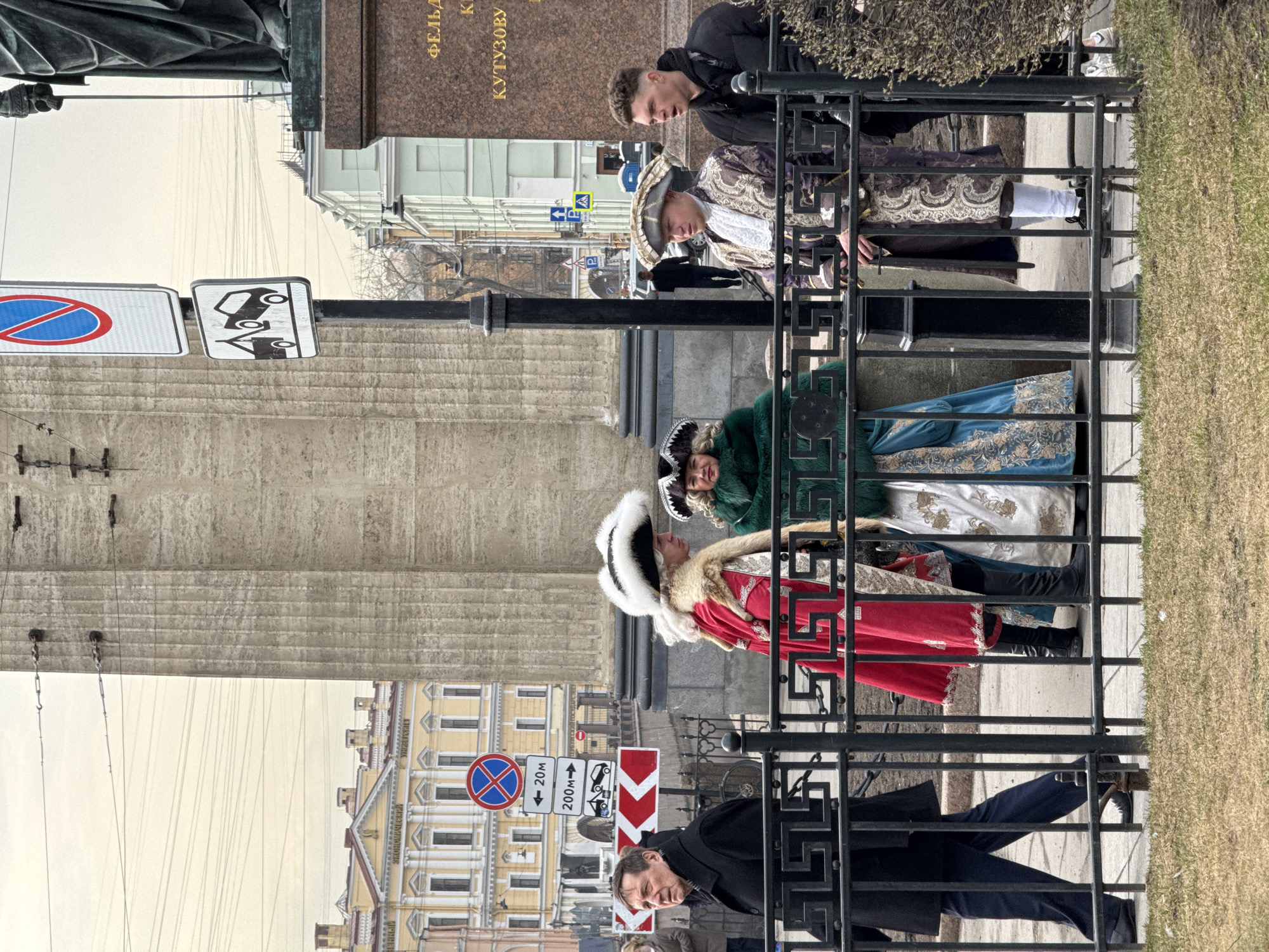 The scene depicts a group of costumed individuals behind a black wrought-iron fence on a street in Saint Petersburg, Russia. The costumes appear to be historical or traditional Russian attire. There are at least four individuals dressed in elaborate outfits, including furs, hats, and embroidered clothing.
Behind the fence, there is a monument or building with the name "ФЕЛЬДМАРШАЛУ КУТУЗОВУ" visible on its base. Street signs are present, including a no-parking sign, a tow-away zone sign, and a sign indicating a 200-meter distance. The architecture in the background is consistent with the style of Saint Petersburg.
Two casually dressed individuals are visible standing on the other side of the costumed group. One is standing over the other. One is standing nearby.
The environment consists of a paved sidewalk, a grassy area behind the fence, and an overcast sky. The overall scene suggests a tourist attraction or a staged photo opportunity.
