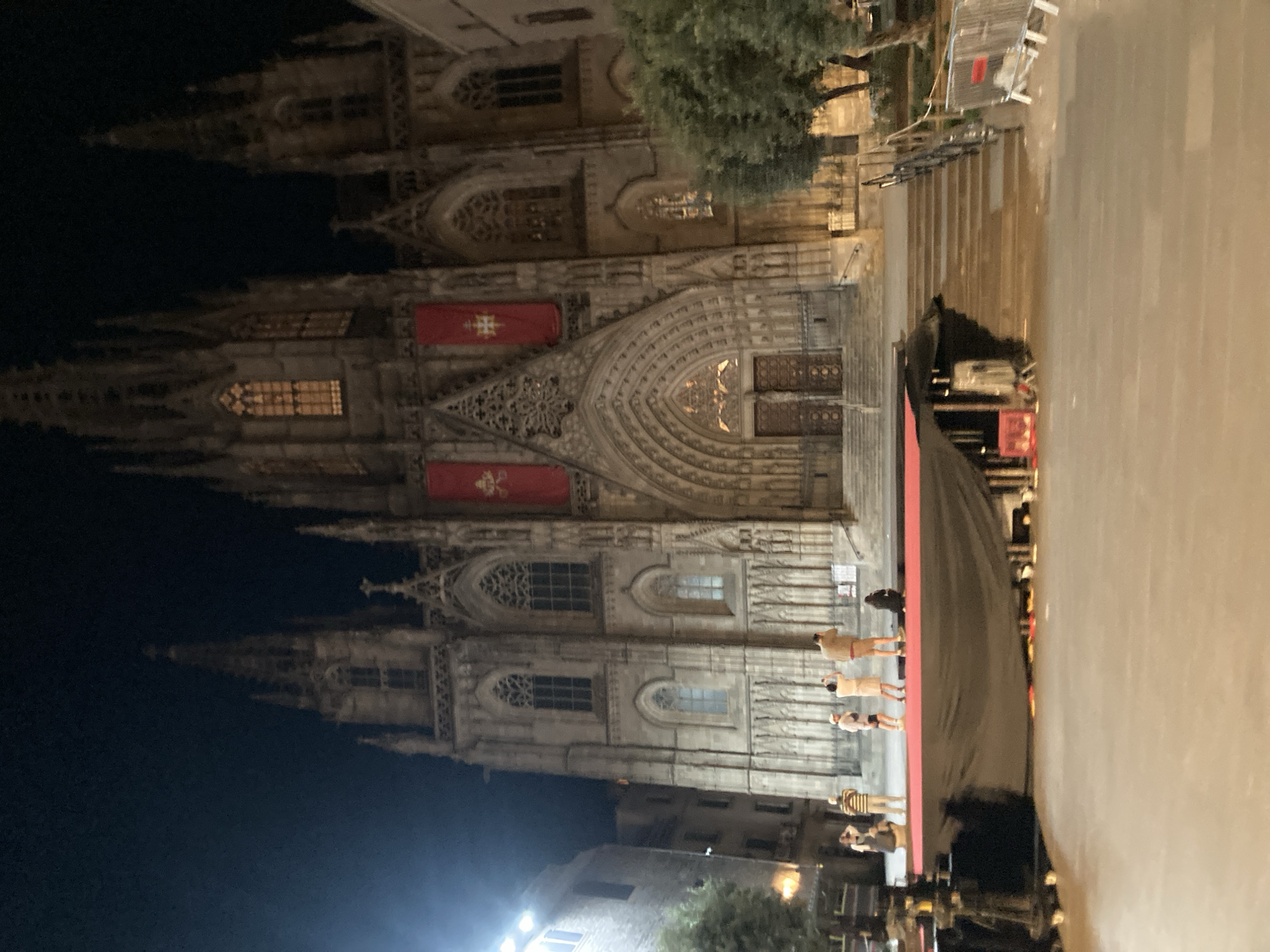 The image captures the illuminated facade of the Barcelona Cathedral (Catedral de la Santa Creu i Santa Eulàlia) at night, located in Barcelona, Spain. In the foreground, a temporary elevated stage structure is present, topped with a red surface, likely a carpet. The base of the stage is covered with dark fabric. To the left of the stage, equipment, possibly lighting or sound apparatus, is partially visible and covered.
Approximately six individuals are standing on the red stage surface, positioned directly in front of the cathedral's main steps. These individuals appear to be observing the cathedral; one is seen holding a device, possibly a camera. Metal crowd control barriers are visible on the right side of the paved plaza.
The cathedral's Gothic facade is prominent, featuring multiple spires, intricate carvings, and numerous arched windows, some of which are illuminated from within, emitting a warm light. Two large rectangular red banners, each displaying distinct ecclesiastical symbols, are prominently hung on the central upper section of the facade, flanking the ornate main entrance. The main entrance consists of a deeply recessed archway with large wooden double doors. Steps ascend from the plaza level to the cathedral entrance. Adjacent buildings are visible to the left of the cathedral, and trees are interspersed within the plaza area, some receiving artificial illumination. The dark sky indicates nighttime conditions.