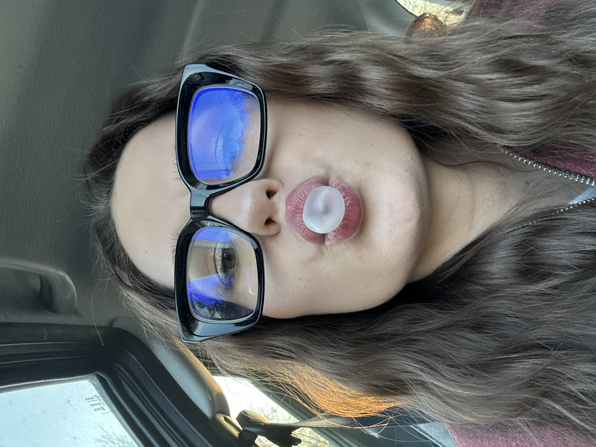 A woman is seen close-up, likely in a vehicle, blowing a bubble gum. She wears black framed glasses. She has long, brown hair. A vehicle interior, including a car door and window, are visible in the lower left corner. The location in Garevaya, Russia cannot be determined from the image.