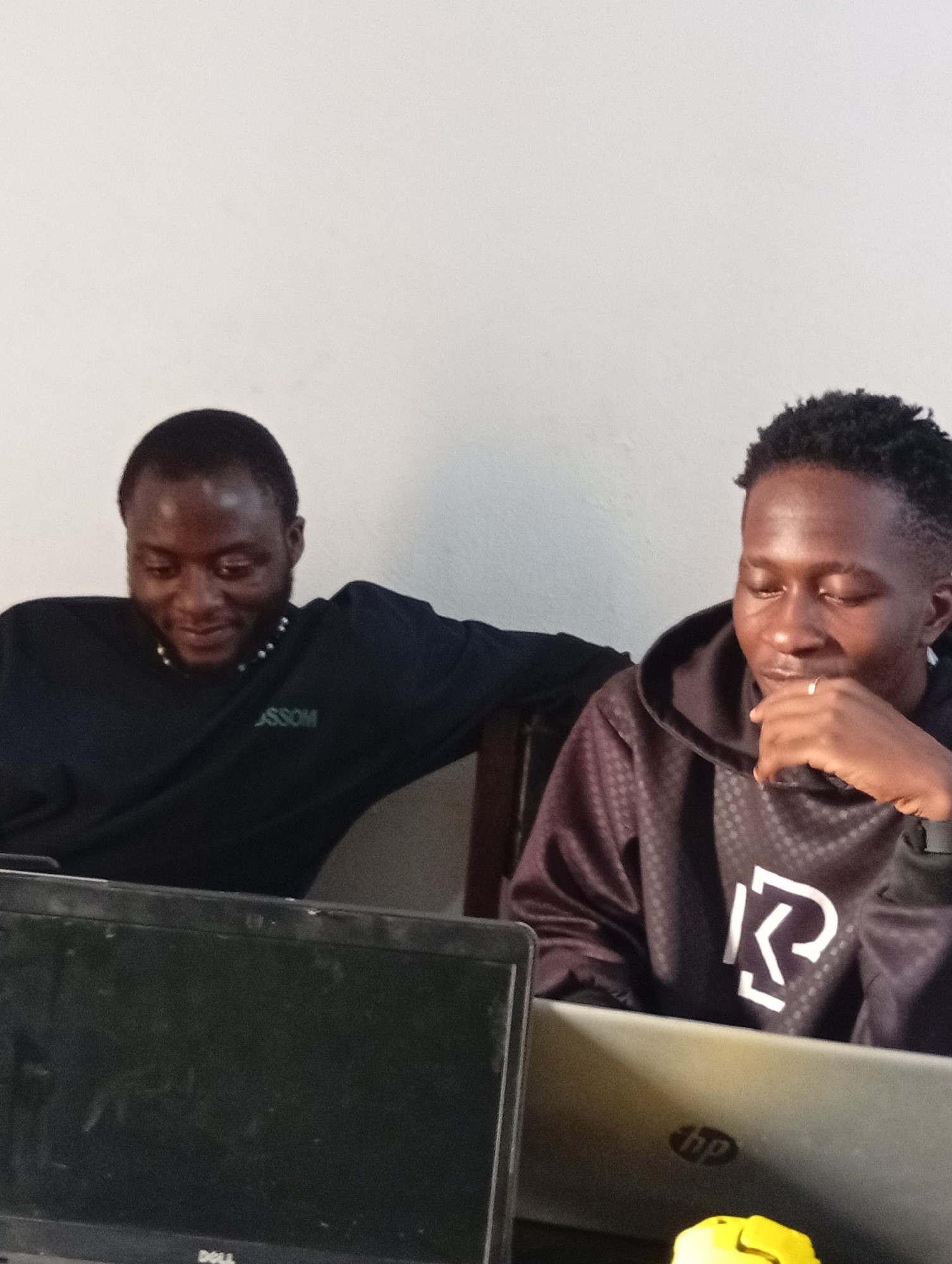 Two young Black men are seated in front of laptops in an indoor setting. The man on the left, with a short haircut and a beard, is wearing a dark long-sleeved shirt with a beaded necklace and a logo that appears to say "JJSSOM". He is looking down and smiling. The man on the right has a fade haircut and is wearing a dark hooded sweatshirt with a white graphic logo on the chest. He is looking down and appears to be deep in thought, with his hand on his chin. In the foreground, a black Dell laptop is visible on the left, and a silver HP laptop is on the right. The background is a plain white wall. The lighting suggests it is daytime, and there are no obvious weather cues. The overall scene appears to be casual and focused on work or study.