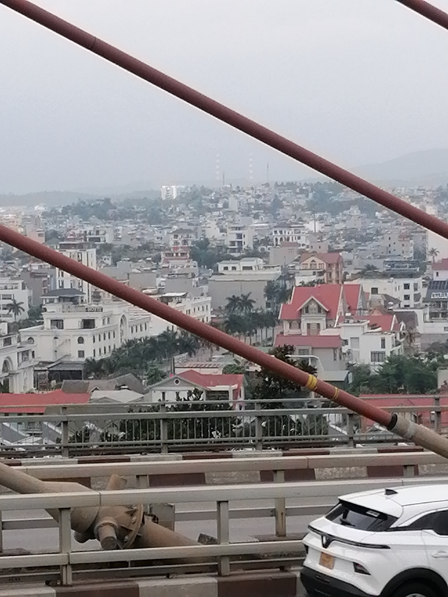 The image presents an urban landscape of Hong Gai Ward, Vietnam, viewed from a bridge on an overcast day. Two prominent, thick red diagonal cables, part of the bridge's support structure, frame the view, extending from the top left to the middle right, and from the middle left to the bottom right.
In the foreground, parts of the bridge deck, railings, and what appears to be a large, rusted pipe or structural component are visible. A white SUV, identifiable as a VinFast vehicle by its distinctive 'V' logo on the rear hatch, is partially seen on the right, driving across the bridge. Its yellow license plate is present but unreadable.
Beyond the bridge structure, a densely built-up city unfolds. Numerous multi-story buildings, predominantly white or light-colored with some featuring red-tiled roofs, are clustered together. Palm trees and other greenery are scattered throughout the urban fabric. In the hazy distance, hills rise, and several tall industrial smokestacks or utility towers are visible against the cloudy sky, suggesting industrial activity in the area. The lighting is diffused and soft, consistent with an overcast daytime, possibly morning or late afternoon. No individuals are distinctly visible in the scene.