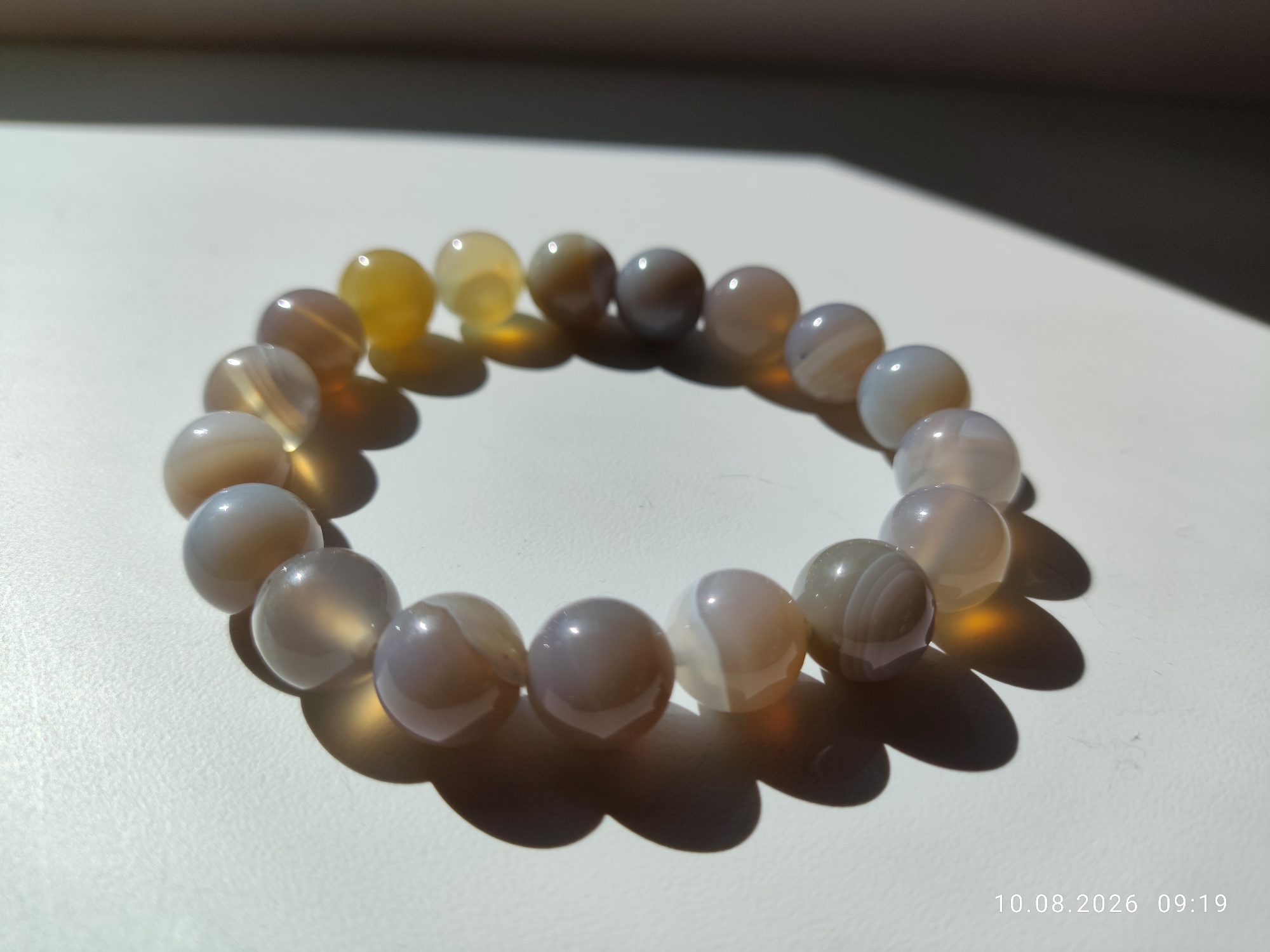 The image shows a bracelet made of semi-transparent, smooth, spherical beads of varying colors, including shades of grey, white, yellow, and amber. The beads are arranged in a circular formation on a white surface. Sunlight casts shadows from the beads onto the surface, creating an oval-shaped shadow for the bracelet. The background is dark and out of focus. In the bottom right corner, a timestamp reads "10.08.2026 09:19". The image does not contain any recognizable landmarks or features that would indicate its location within Pavlodar, Kazakhstan.