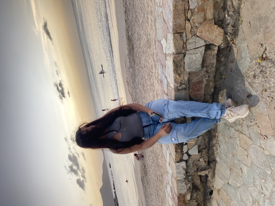The image depicts a coastal scene in Juan Griego, Venezuela, under the lighting conditions of either sunrise or sunset. A young female is seated in the foreground on a low stone wall constructed from irregular, flat stones. She is oriented slightly to her right, wearing a grey sleeveless top, light blue cargo-style jeans with elastic cuffs, and light-colored athletic shoes. Her dark hair partially obscures the right side of her face as she looks towards the right. Her left hand holds a dark-colored smartphone. The stone wall separates a paved ground area, also made of irregular stones, from a sandy beach visible to the right. Beyond the beach, calm ocean water extends towards the horizon. Several individuals are present in the water; one person is on a paddleboard further out, and others are wading closer to the shore. A child is visible kneeling on the sand to the far left. In the distance, mountains or hills and a rocky landmass are visible on the left horizon. The sky displays a gradient of warm yellow and orange tones near the horizon, transitioning to lighter blue and grey overhead.