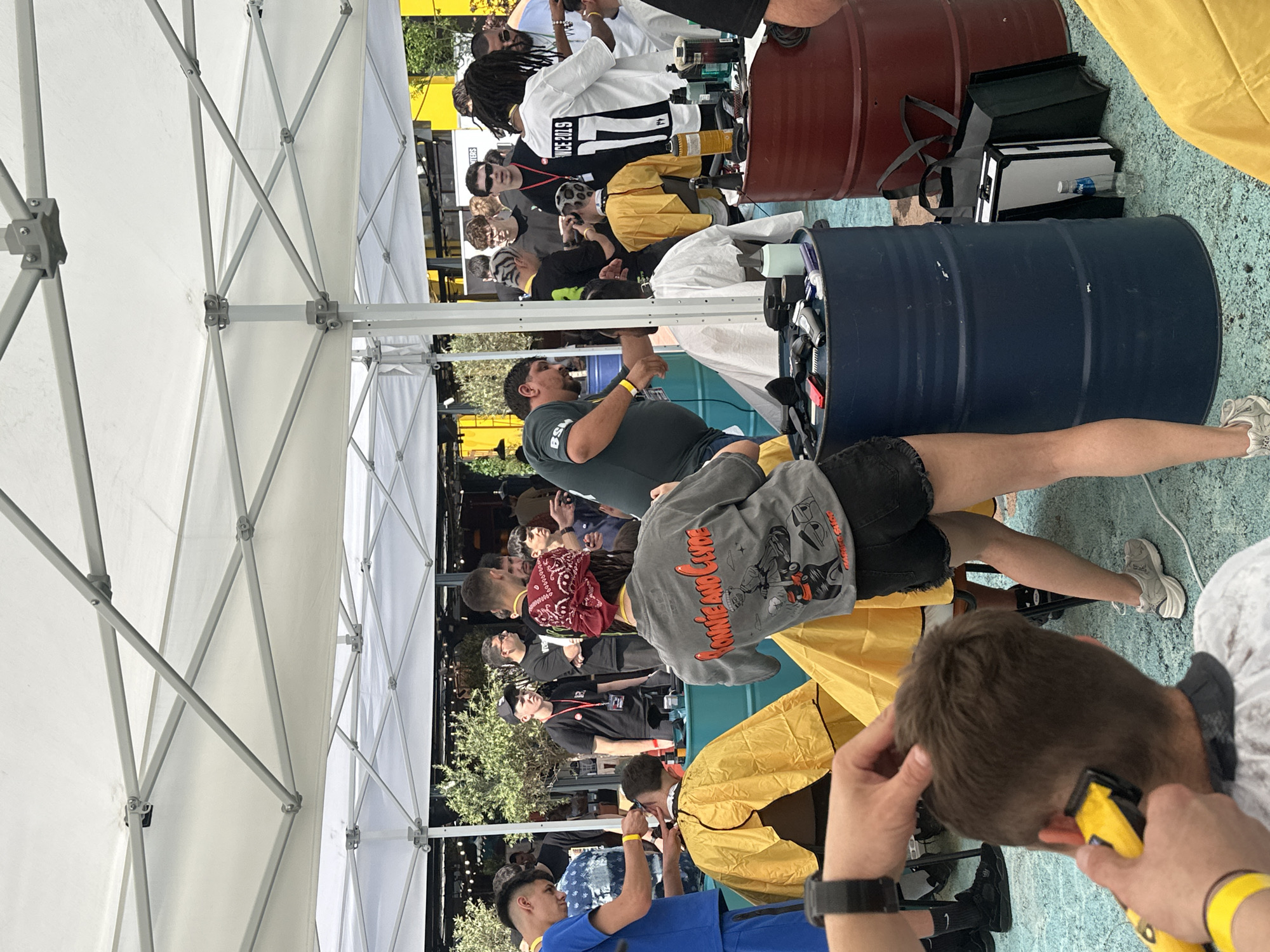This image depicts an outdoor barbering event or competition taking place under several white pop-up canopies in Sofia, Bulgaria. The scene features multiple barber stations, each typically consisting of a barber and a client, with various individuals observing or participating.
In the foreground, a person (partially visible, bottom left) is shown from the waist down, holding a yellow electric hair clipper and actively trimming the back of a male client's head. The client is wearing a white barber cape. To the right, a woman is seen from behind, wearing a grey t-shirt with "BONNIE AND CLYDE" and a graphic of cartoon characters on a motorcycle, black shorts, and a red bandana on her head. She stands near a dark blue metal barrel, which serves as a makeshift workspace holding various barber tools like clippers, spray bottles, and containers.
Further into the midground, several more barber stations are active. On the left, a man in a blue t-shirt and shorts is cutting a client's hair. The client is draped in a yellow barber cape. In the center, a man with distinctive zebra-striped hair is cutting another client's hair, who is also wearing a yellow cape. To the right, a man with dreadlocks, wearing a white jersey with "SINCE 2019 11" on the back, is attending to a client. Another barber in a dark green t-shirt with "BSM" on the shoulder is visible on the far left, also cutting hair.
The barber stations utilize large metal barrels, both blue and red, as surfaces for tools and equipment. On the ground near the blue barrel, a clear water bottle is visible next to a black bag and a white and silver case, possibly containing barber supplies. The ground is a light blue, textured surface, possibly a mat or painted concrete.
In the background, more people are visible, including individuals standing and observing. Structures with yellow and black elements, possibly buildings or kiosks, are present, along with green foliage and string lights, suggesting a festive or commercial outdoor setting. The overall lighting indicates it is daytime.