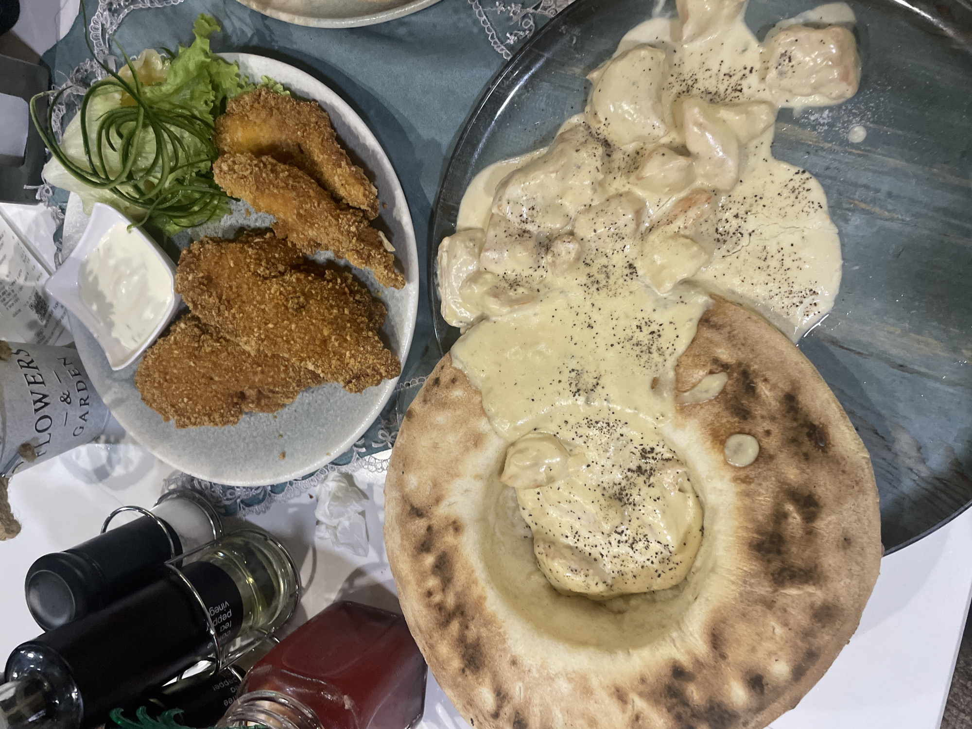 The image shows two plates of food on a table. On the left, a white plate contains fried chicken, lettuce, cucumber spirals, and a small bowl of white sauce. On the right, a dark grey plate holds a round loaf of bread filled with a creamy sauce and meat. Spice bottles and a small jar are visible in the lower left corner. Based on the food presentation style, the location could be a restaurant or home in Gabrovo, Bulgaria, though definitive confirmation of the city and country is not possible from the visual data alone. There are no people visible in the image.